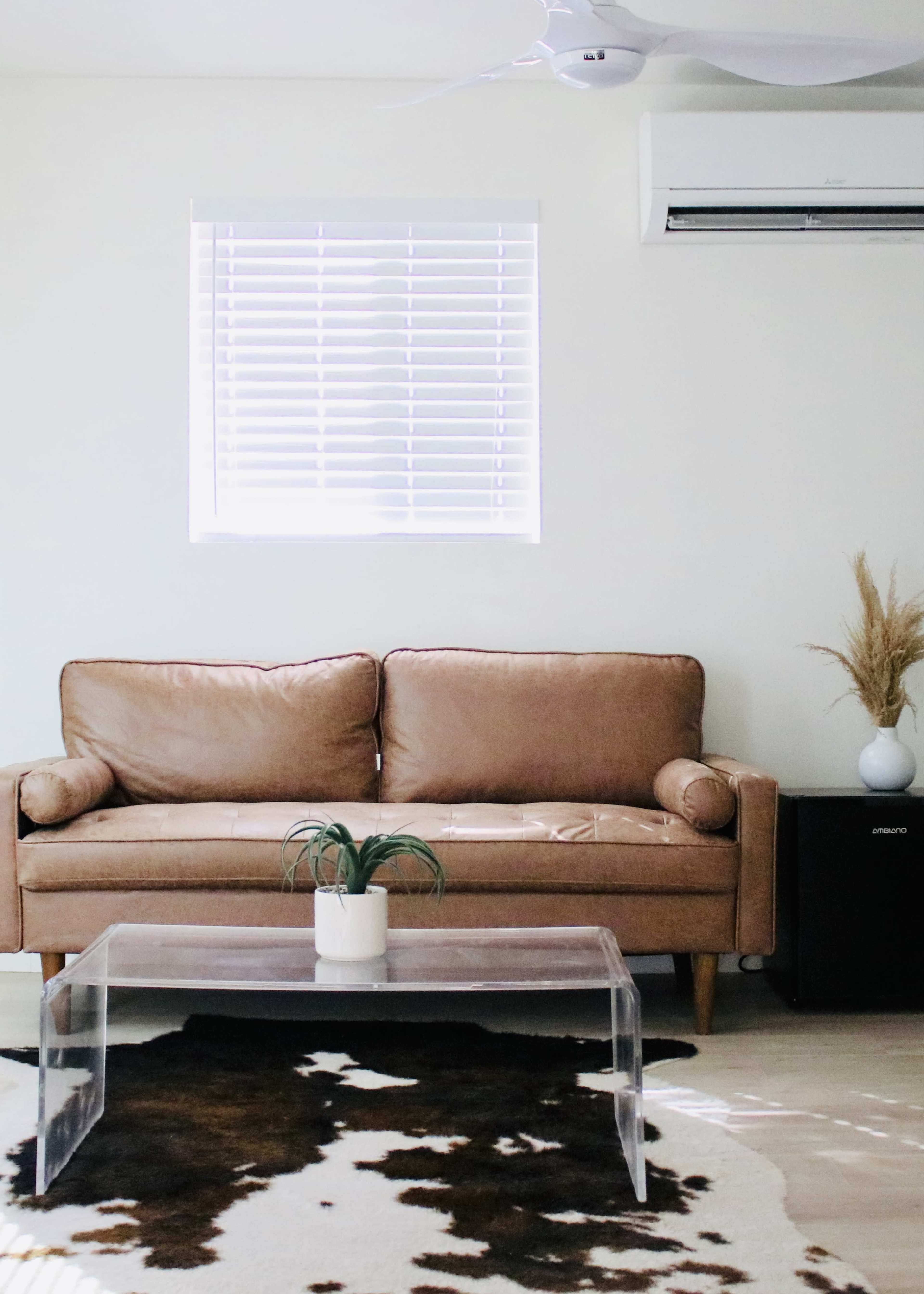 A leather sofa is positioned opposite a transparent coffee table on a cowhide rug, with a small plant and a decorative vase nearby against a light-colored wall.