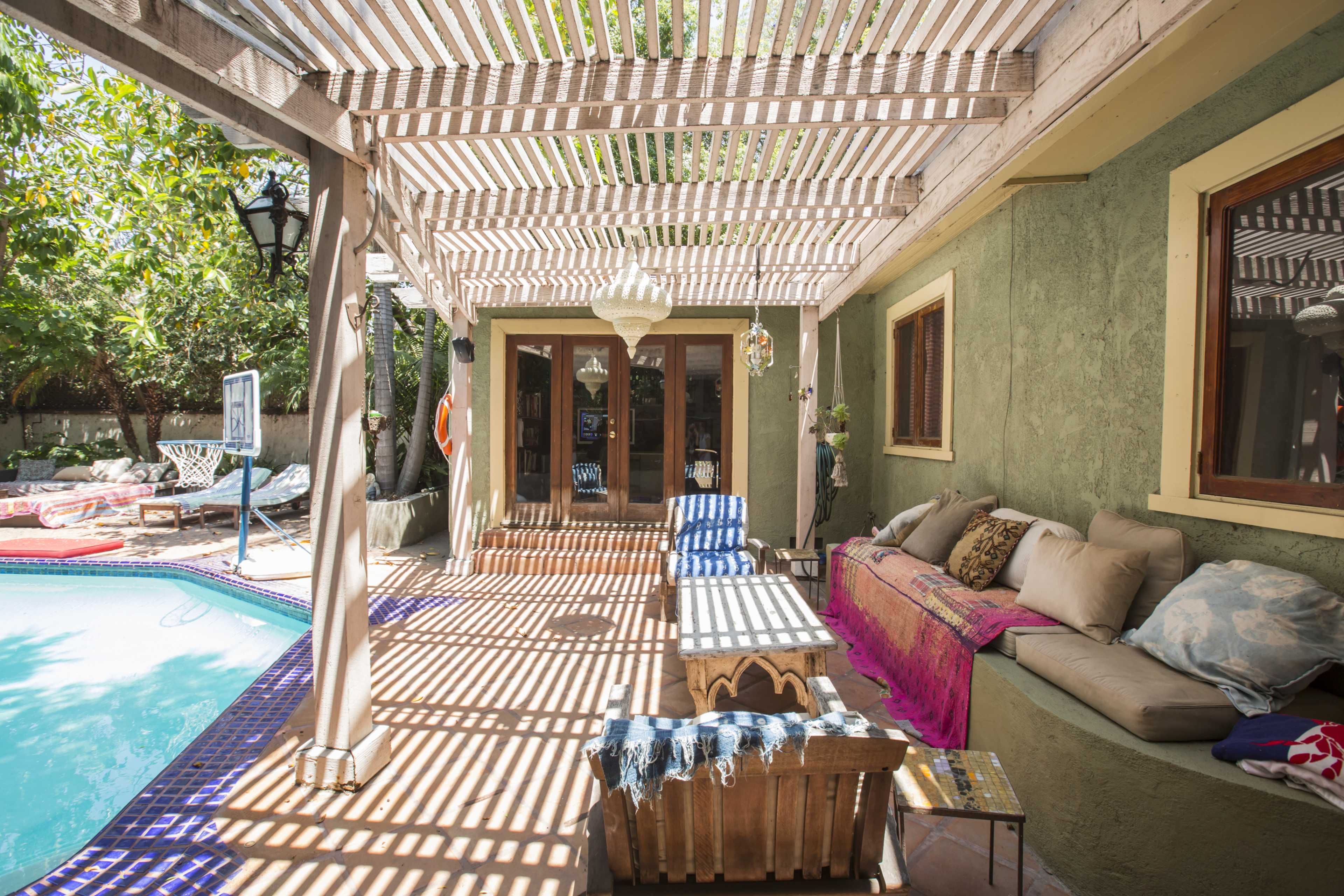 The image shows a patio area with a wooden structure overhead, a pool adjacent to it, and seating arranged along the side.