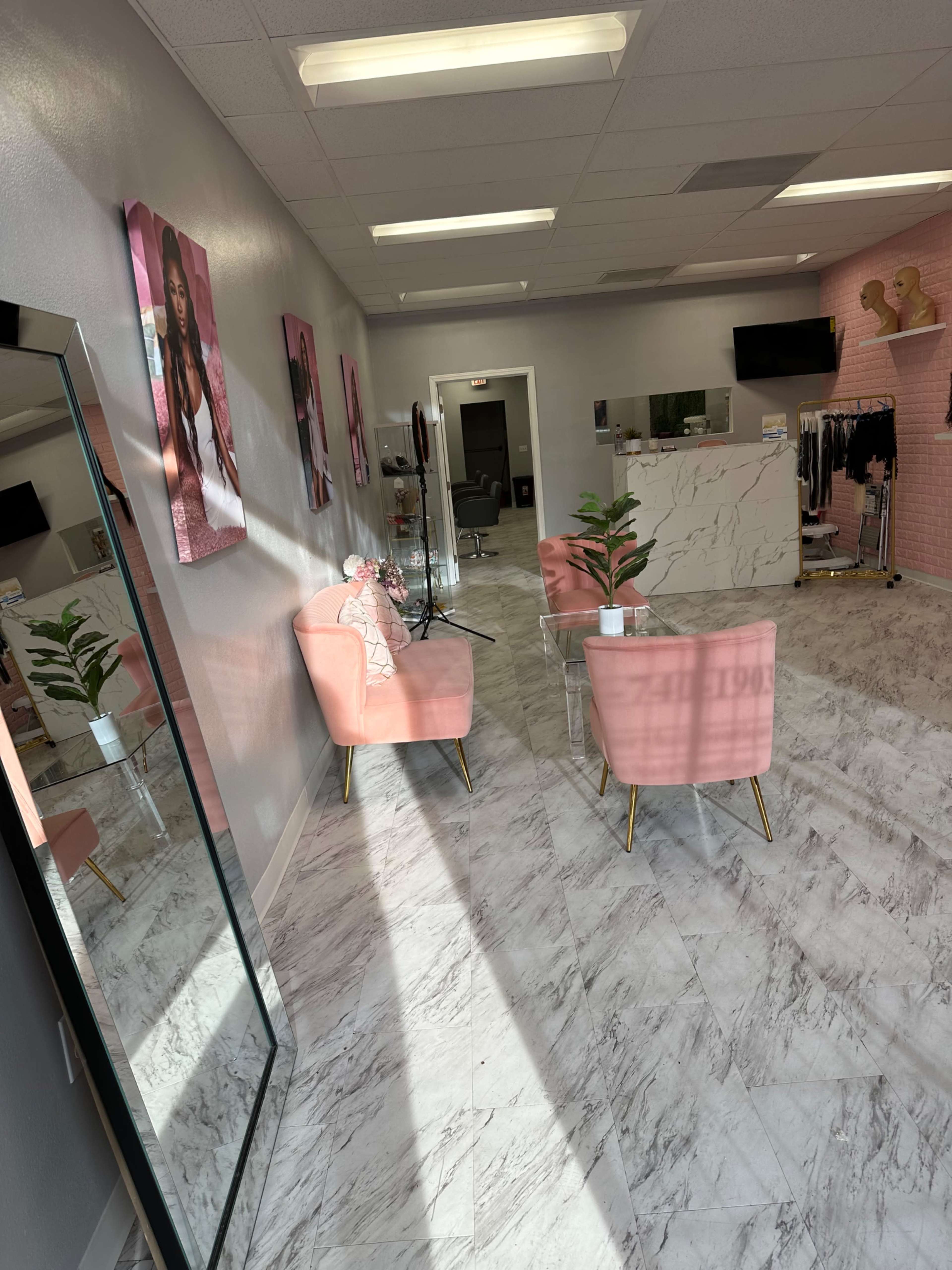 A modern salon interior with pink chairs, a coffee table, plants, and images on the walls, featuring a mirrored wall and styling stations in the background.
