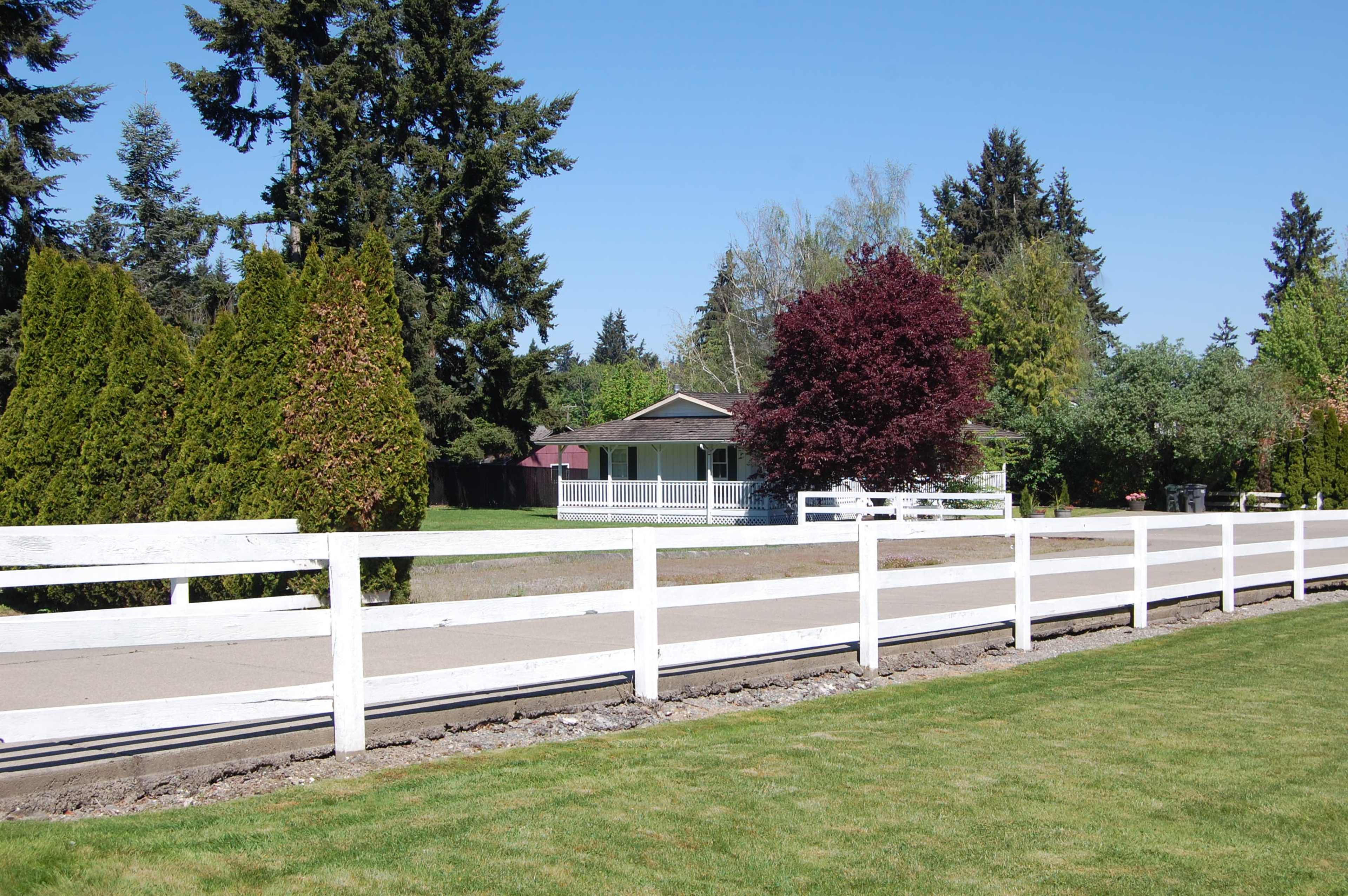 A white fence lines a grassy area in front of a single-story house surrounded by trees and shrubs.