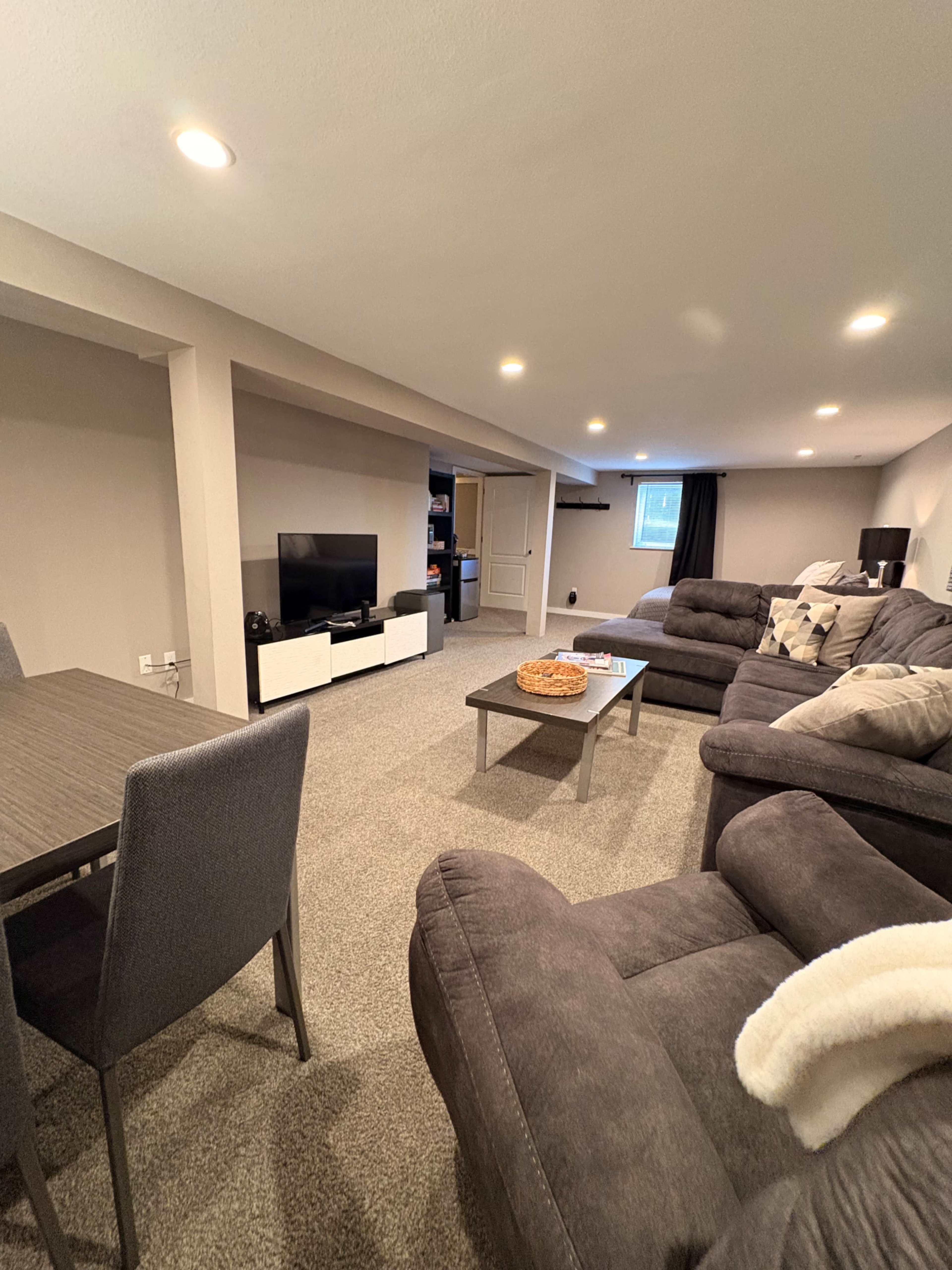 The image shows a cozy living space with gray carpet, a sectional sofa, a coffee table, a dining area, and a television on a white console.