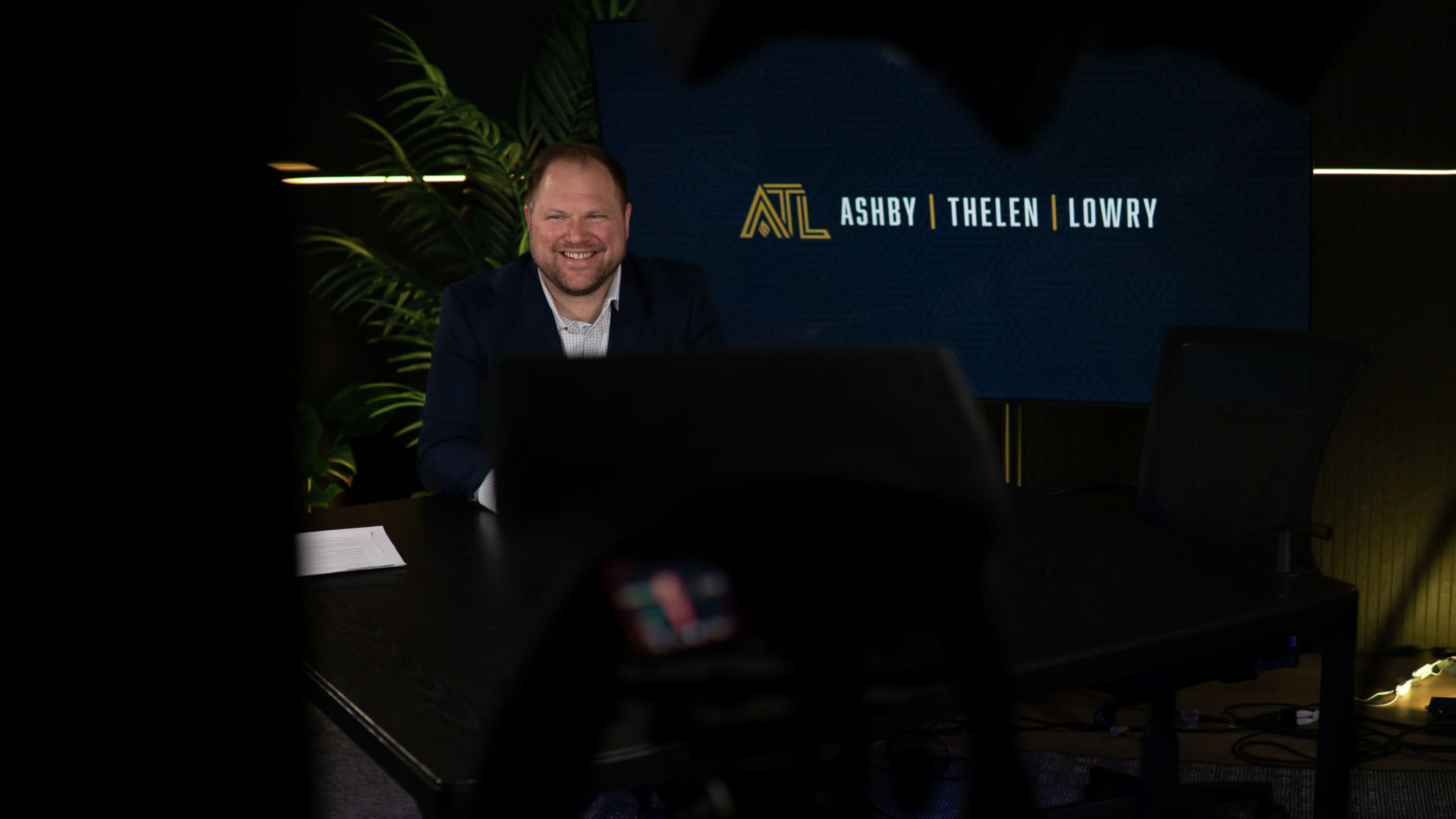 A man sits at a desk, smiling, in front of a backdrop featuring the names "Ashby | Thelen | Lowry."