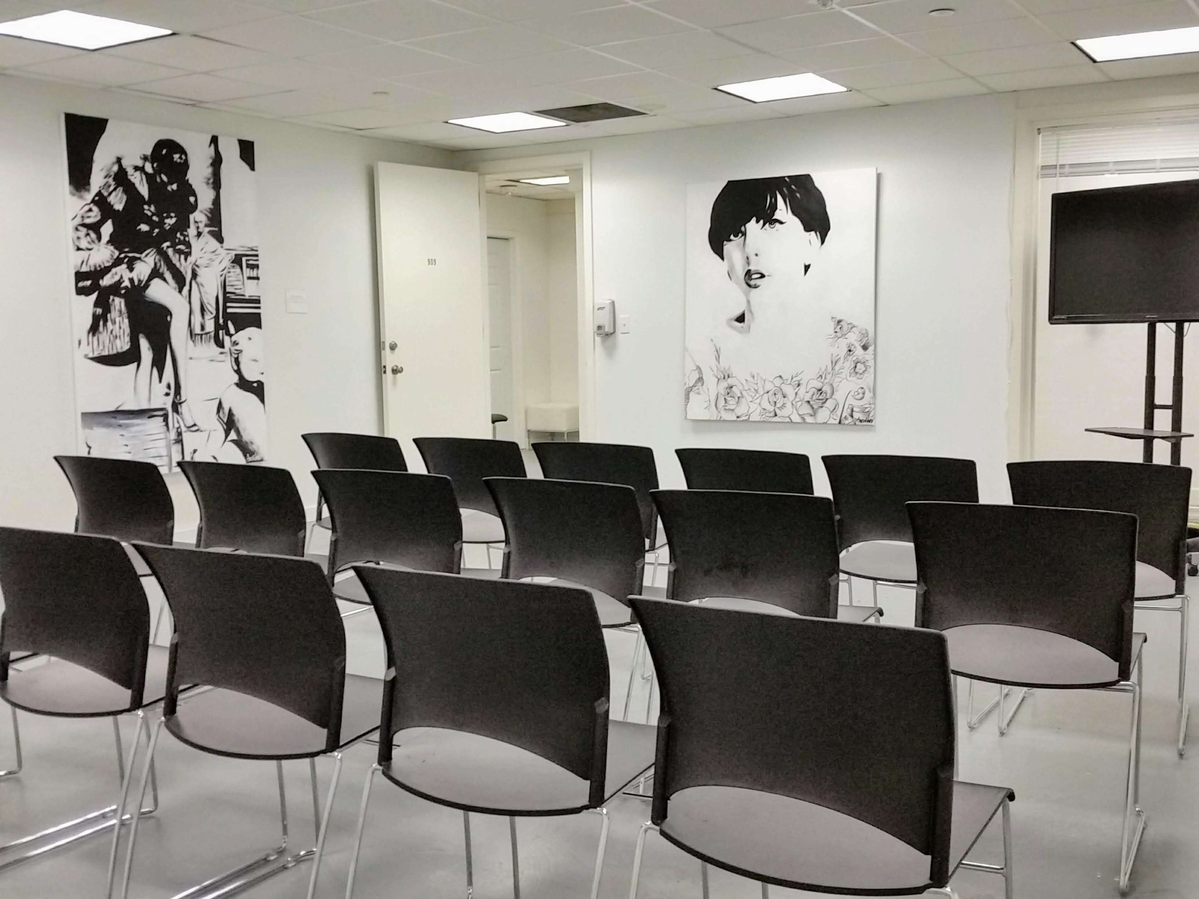 A sparsely furnished room with black chairs set in rows, featuring artwork on the walls and a television screen in the corner.