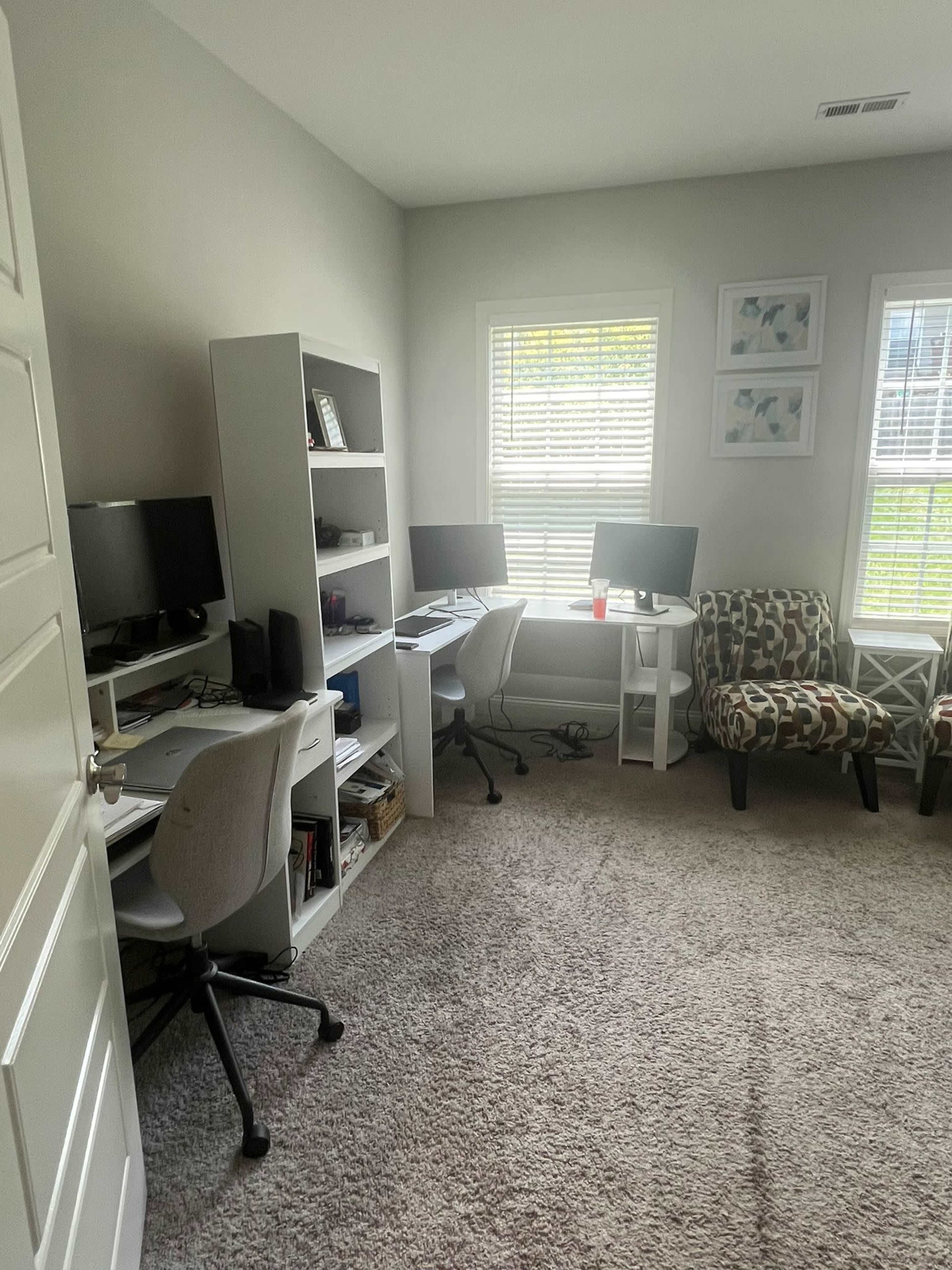 A home office with two desk setups, bookshelves, and patterned chairs, all situated in a softly lit room with windows.