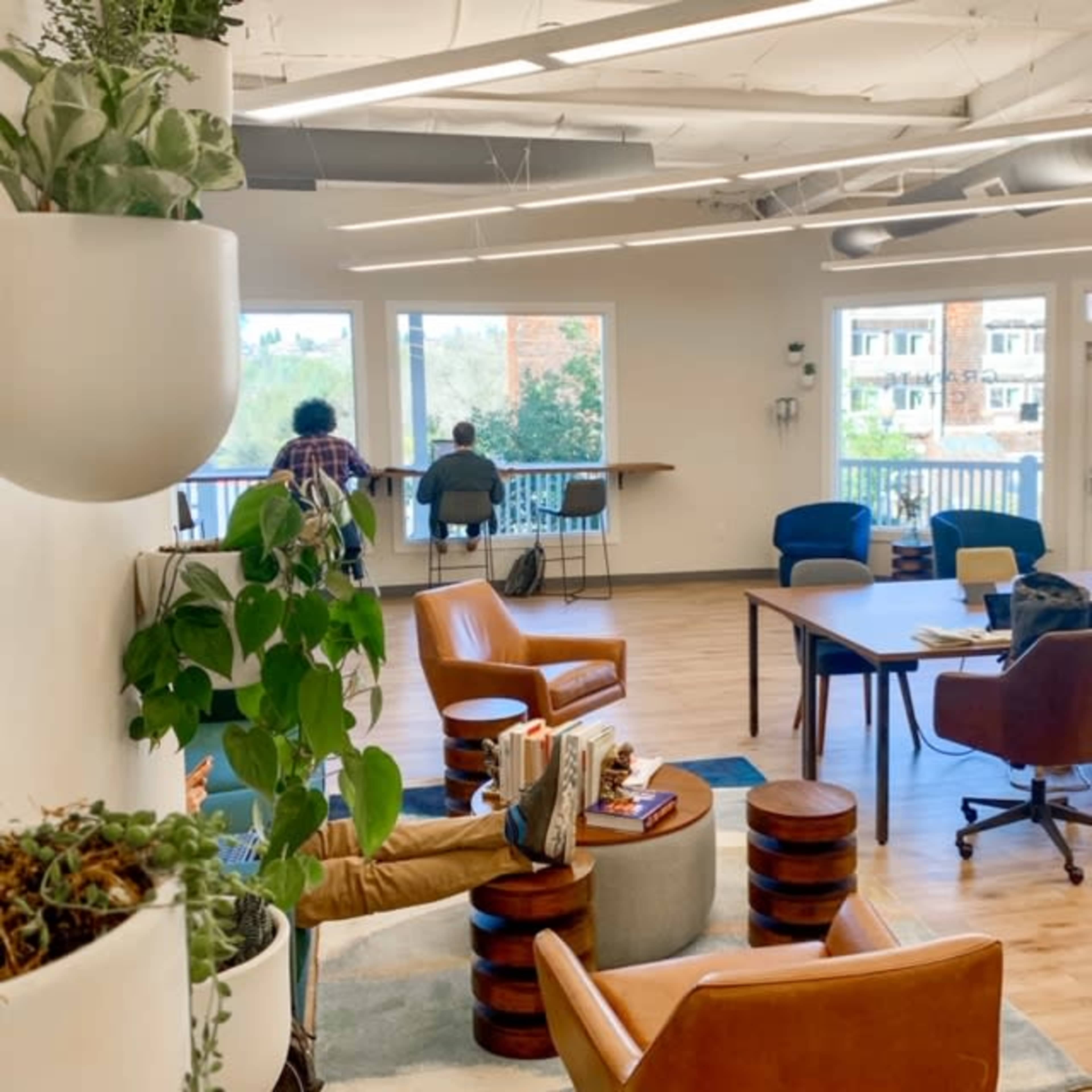 The image depicts a modern office space with plants, cozy seating, and a view of the outdoors through large windows.