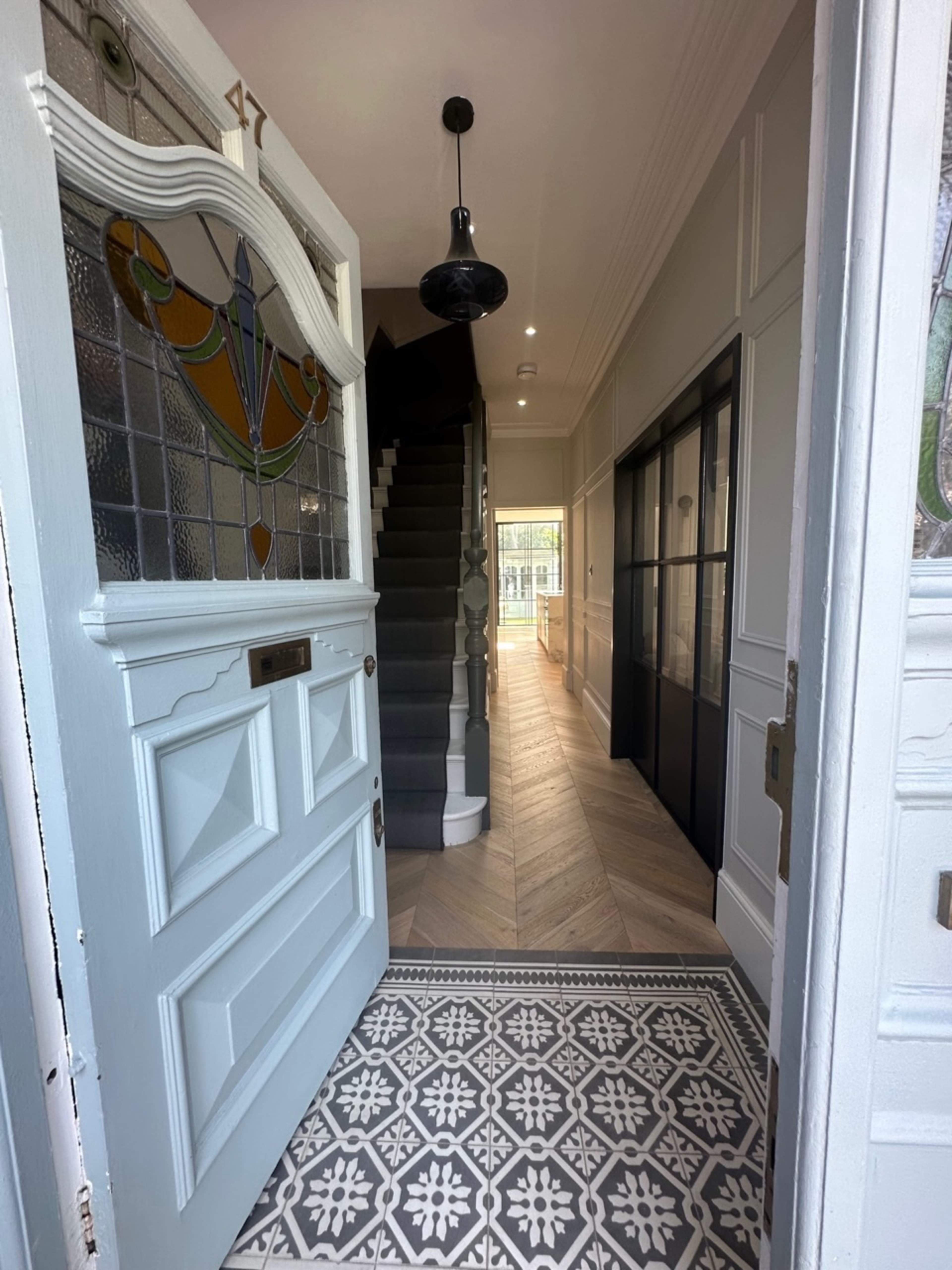 The image shows an open front door revealing a bright hallway with a staircase, patterned tile flooring, and a view of a glass-paneled door at the end.