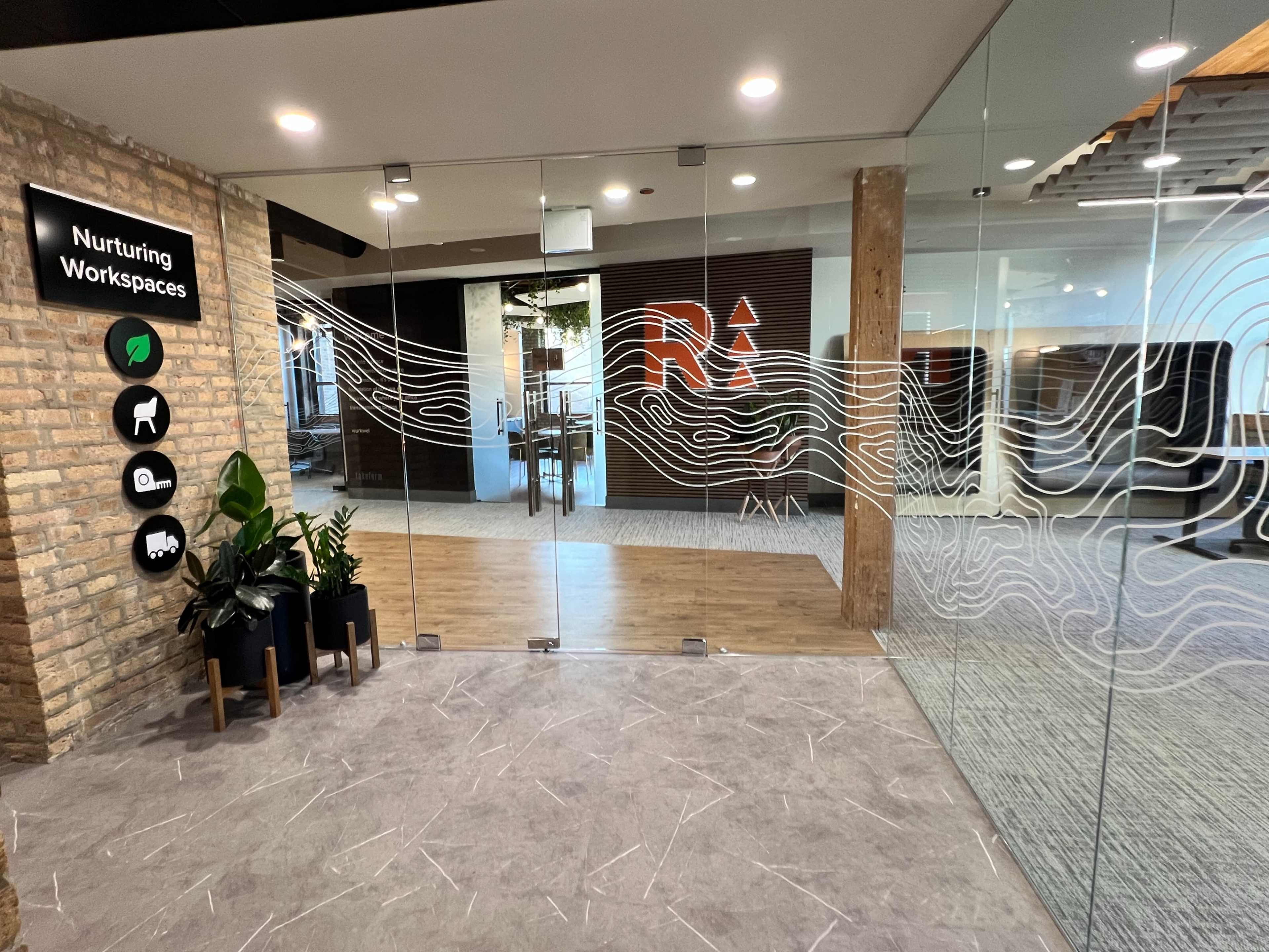 The image shows a glass entrance to an office space, featuring decorative wave patterns and a sign indicating "Nurturing Workspaces."