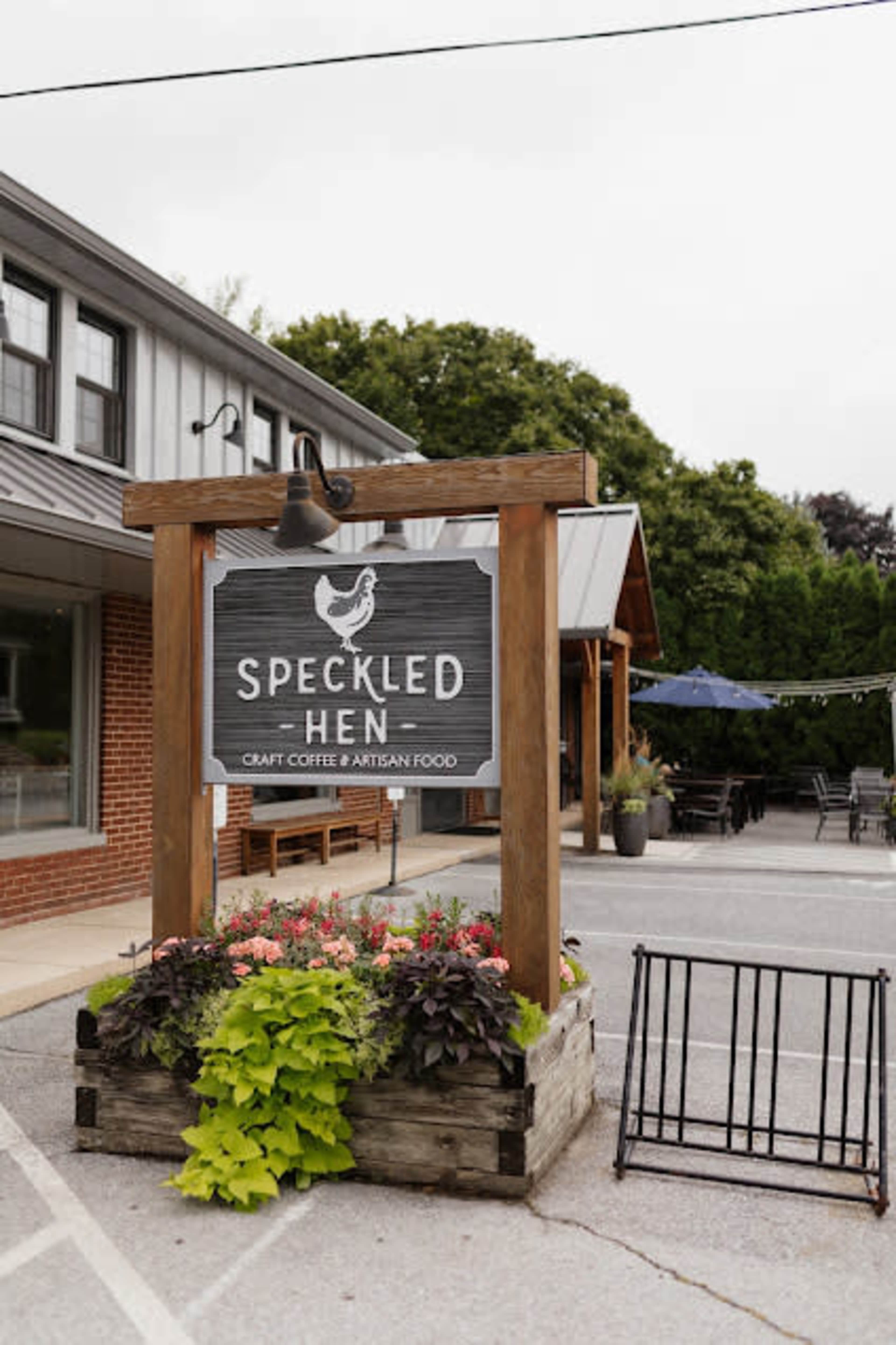 The image shows a sign for "Speckled Hen," a café offering craft coffee and artisan food, set in a landscaped planter at the entrance of a building.