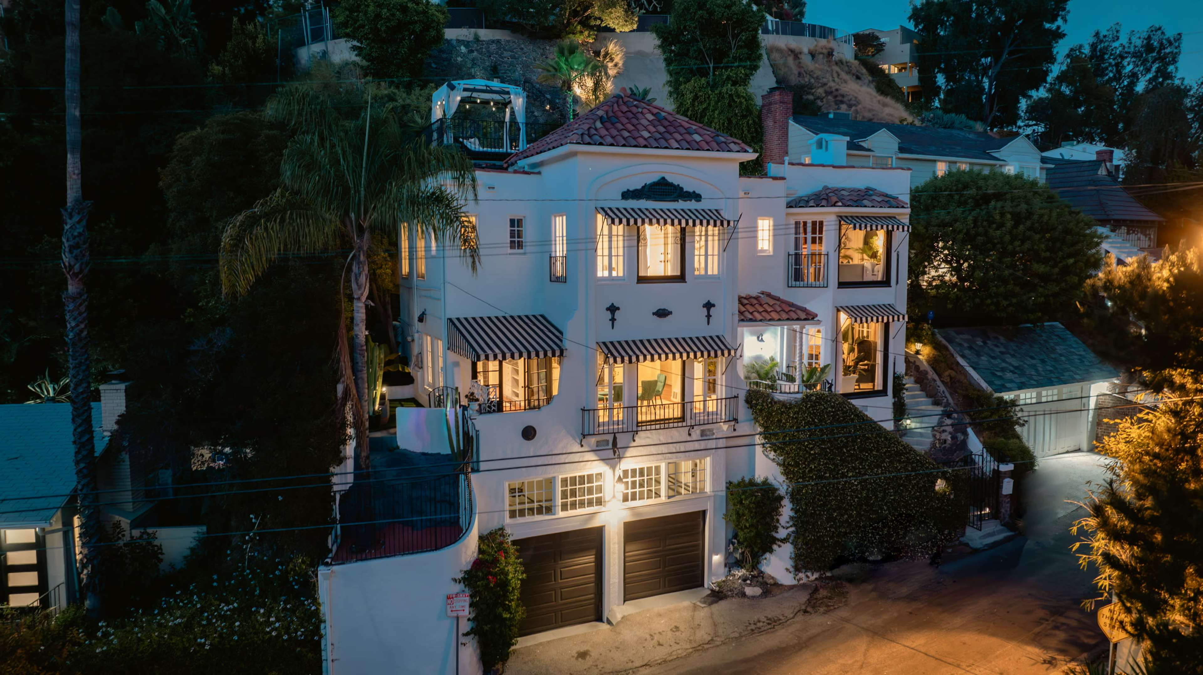 The image shows a large, two-story white house with a terracotta roof, illuminated at night, located on a hillside with surrounding vegetation.