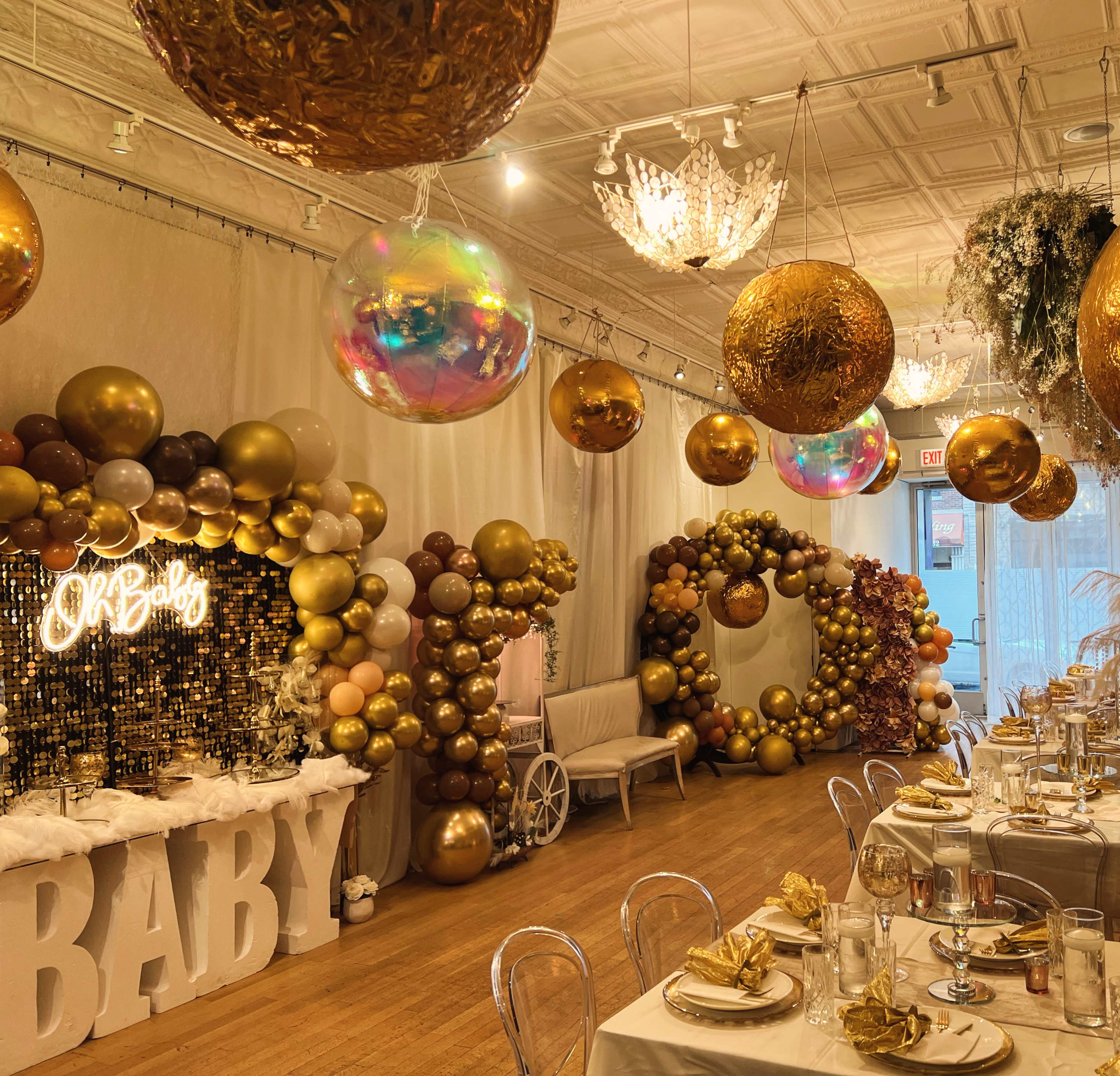 The image depicts a decorated event space with gold and white balloons, elegant table settings, and a "Baby" sign illuminated in lights.