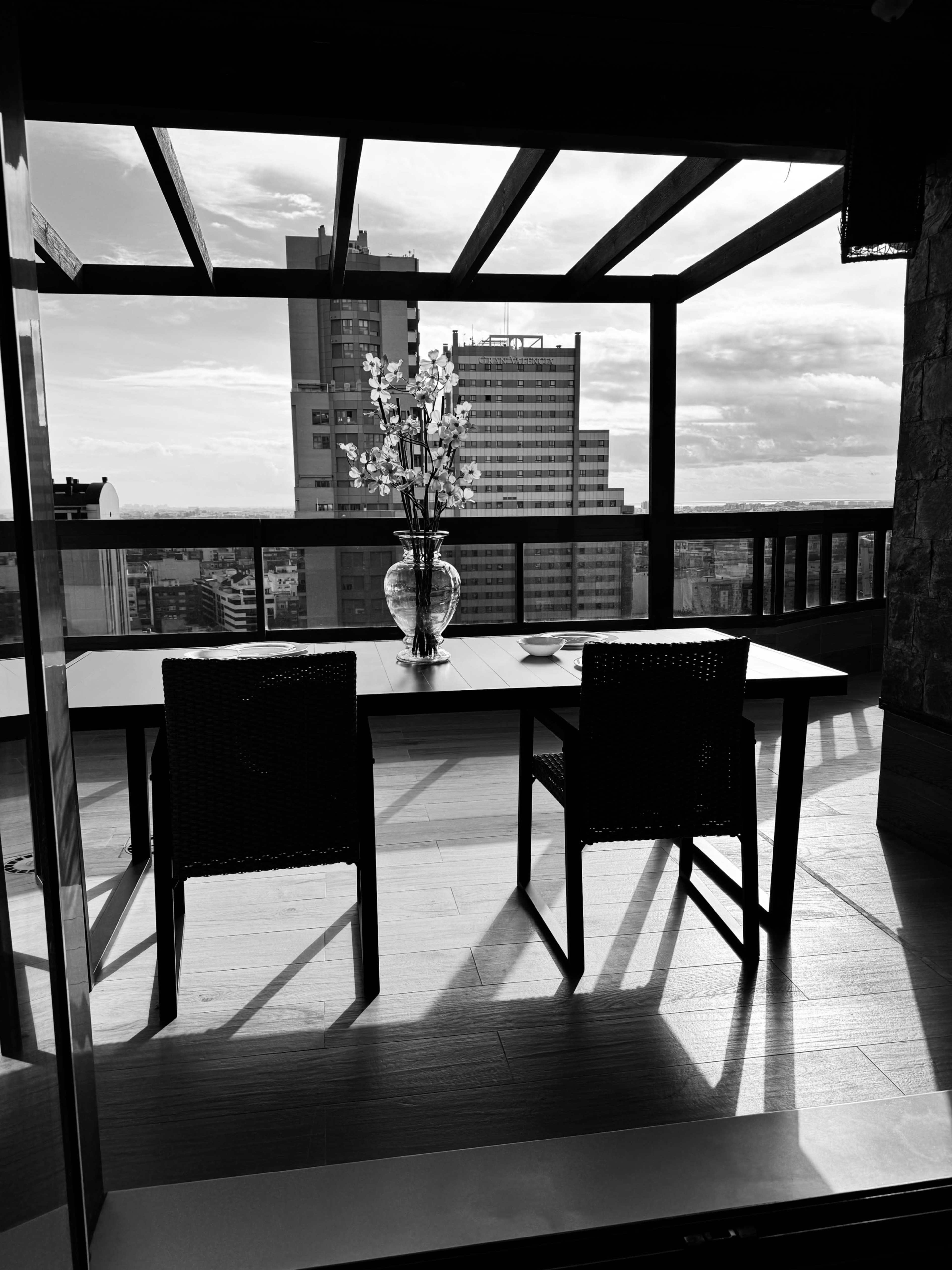 The image depicts a minimalist dining area with two chairs and a vase of flowers, overlooking a city skyline through large glass doors.