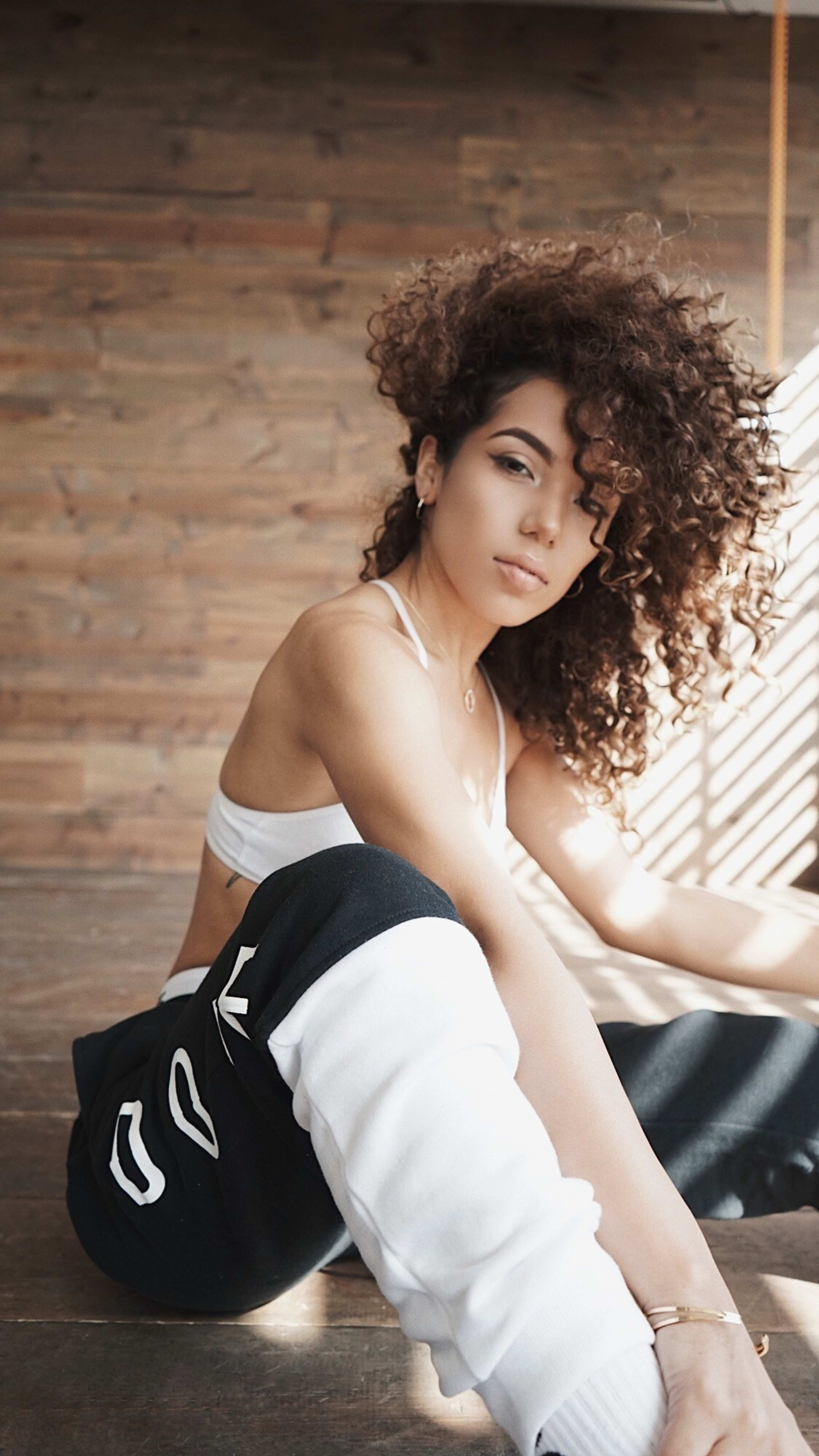 A young woman with curly hair sits on a wooden floor, wearing a white crop top and black sweatpants, with sunlight filtering through blinds in the background.