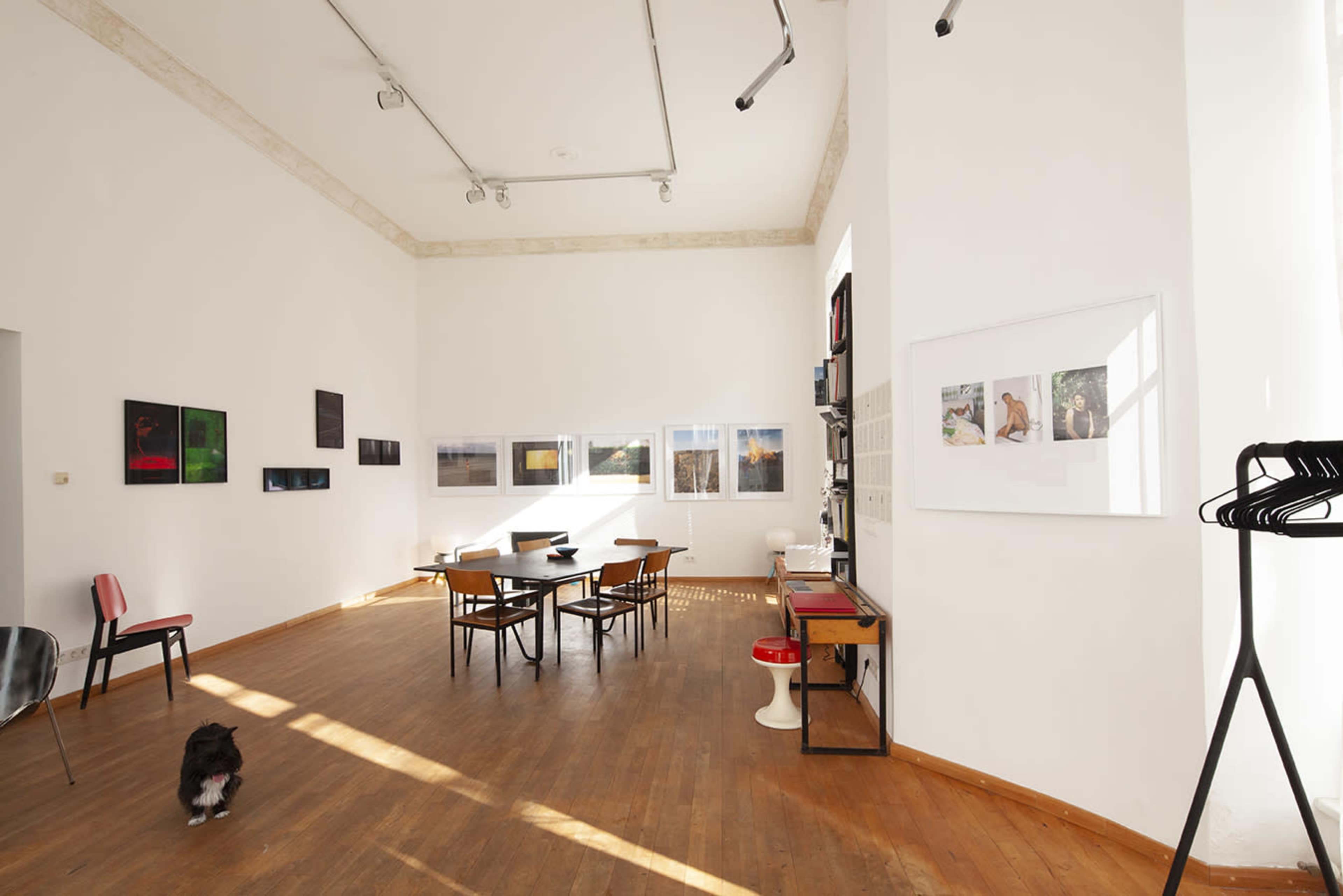An art gallery with wooden floors, a central table surrounded by chairs, and photographs displayed on the walls.