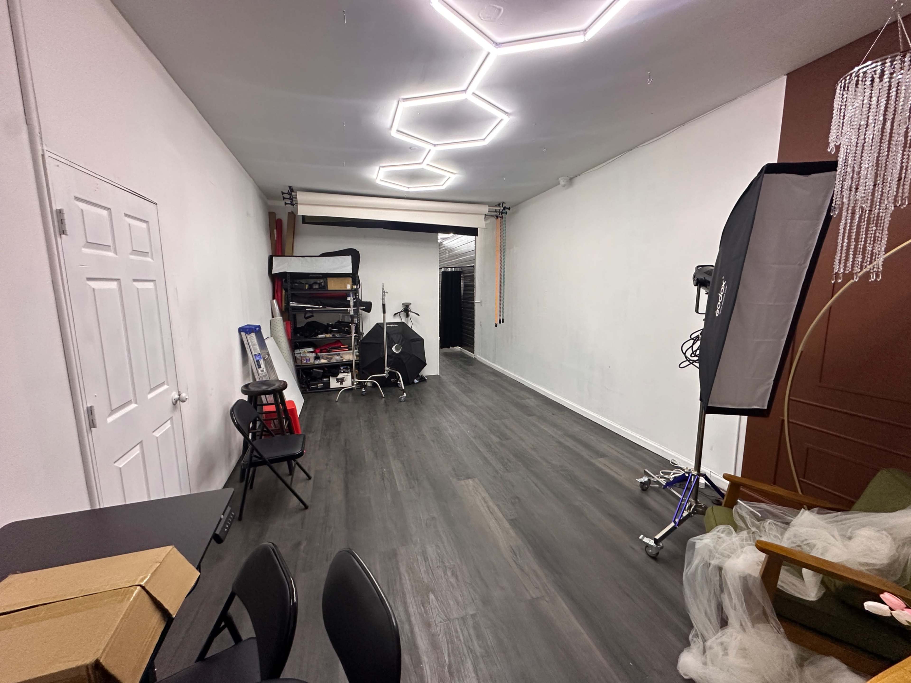 The image shows a spacious room with hardwood flooring, featuring photography equipment on one side and a chair in the corner.