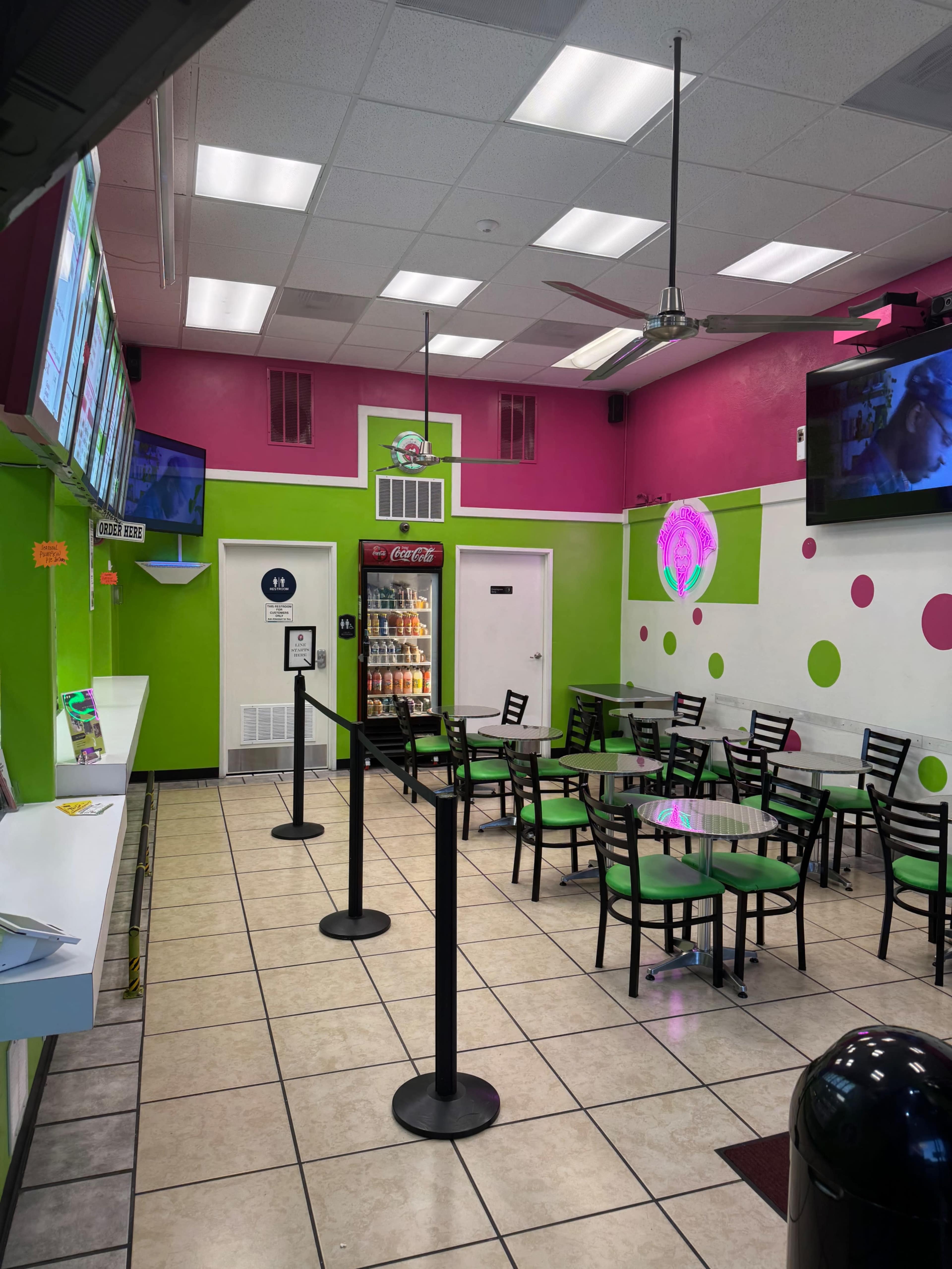 The image shows a colorful fast-food restaurant interior with green and pink walls, a seating area featuring black chairs and glass tables, and a display of drinks in a cooler.