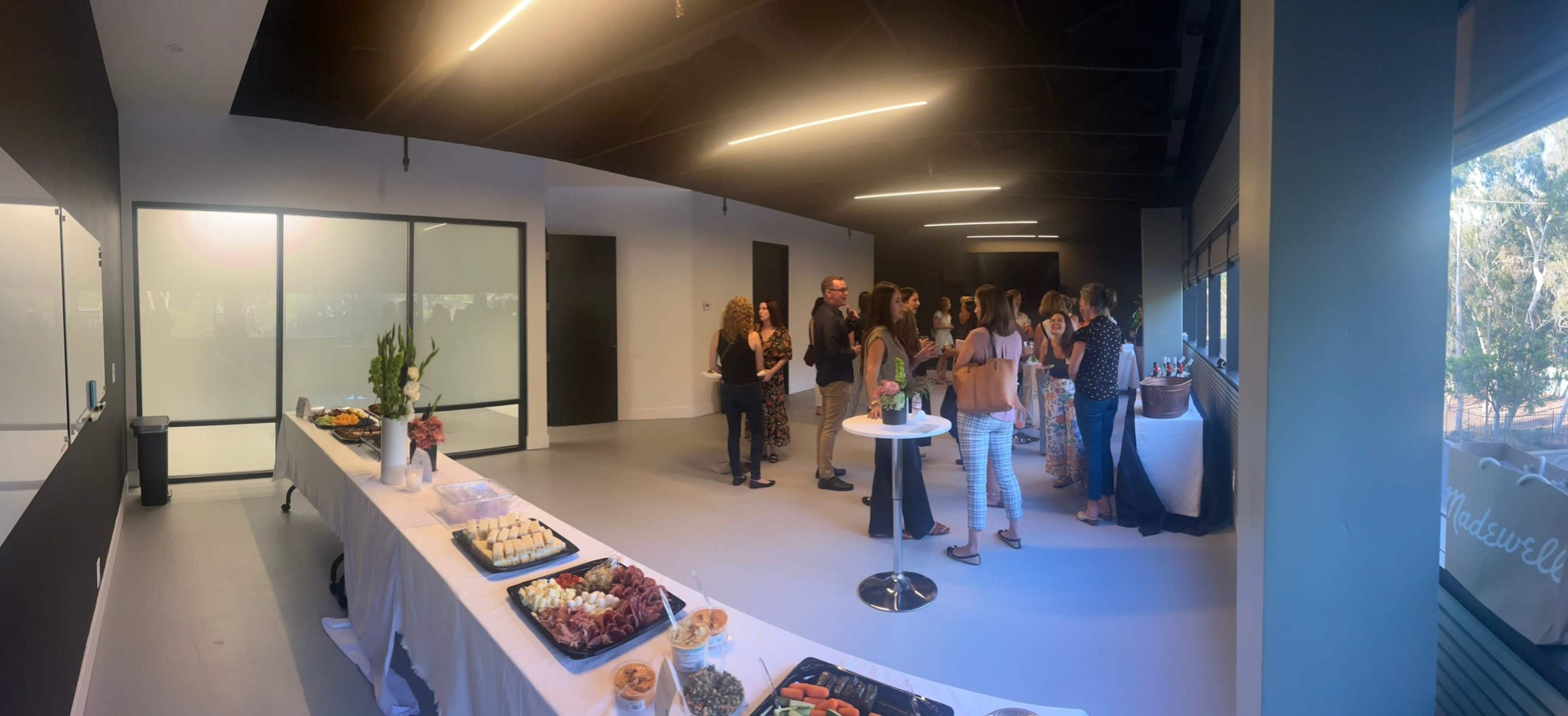 A group of people is gathered in a spacious event room with a table displaying various appetizers and a large window providing natural light.