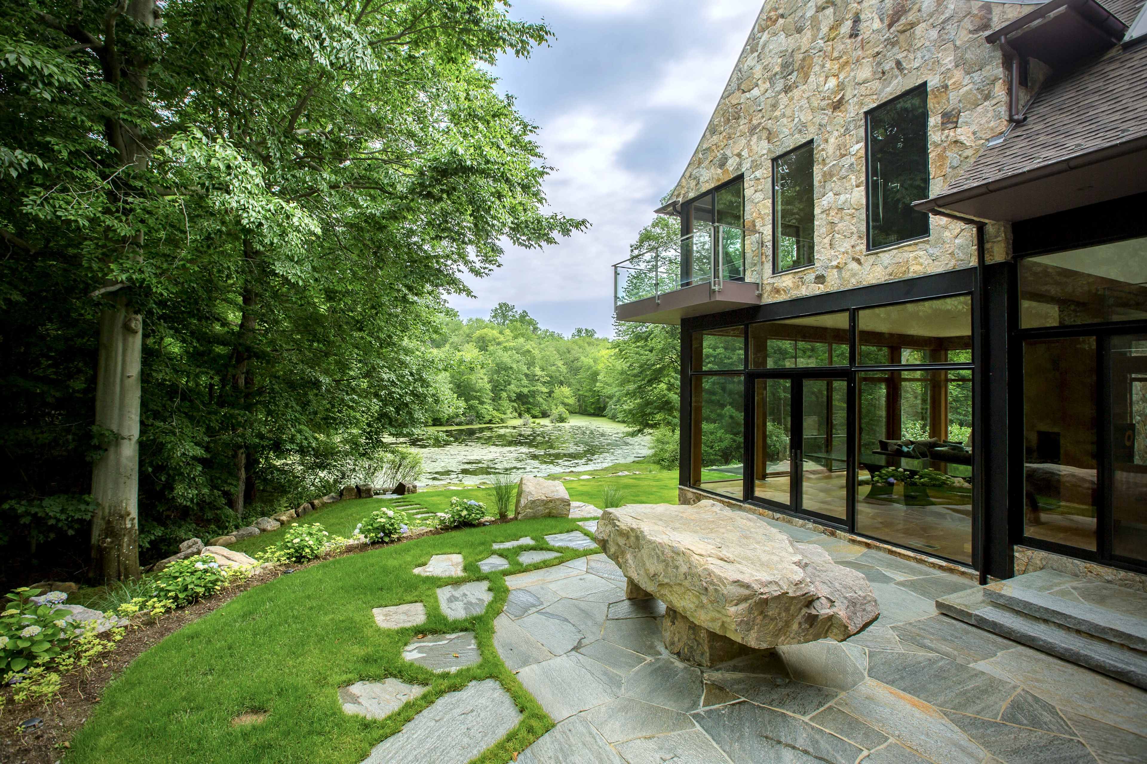 The image shows a modern stone house with large glass windows overlooking a pond surrounded by greenery.