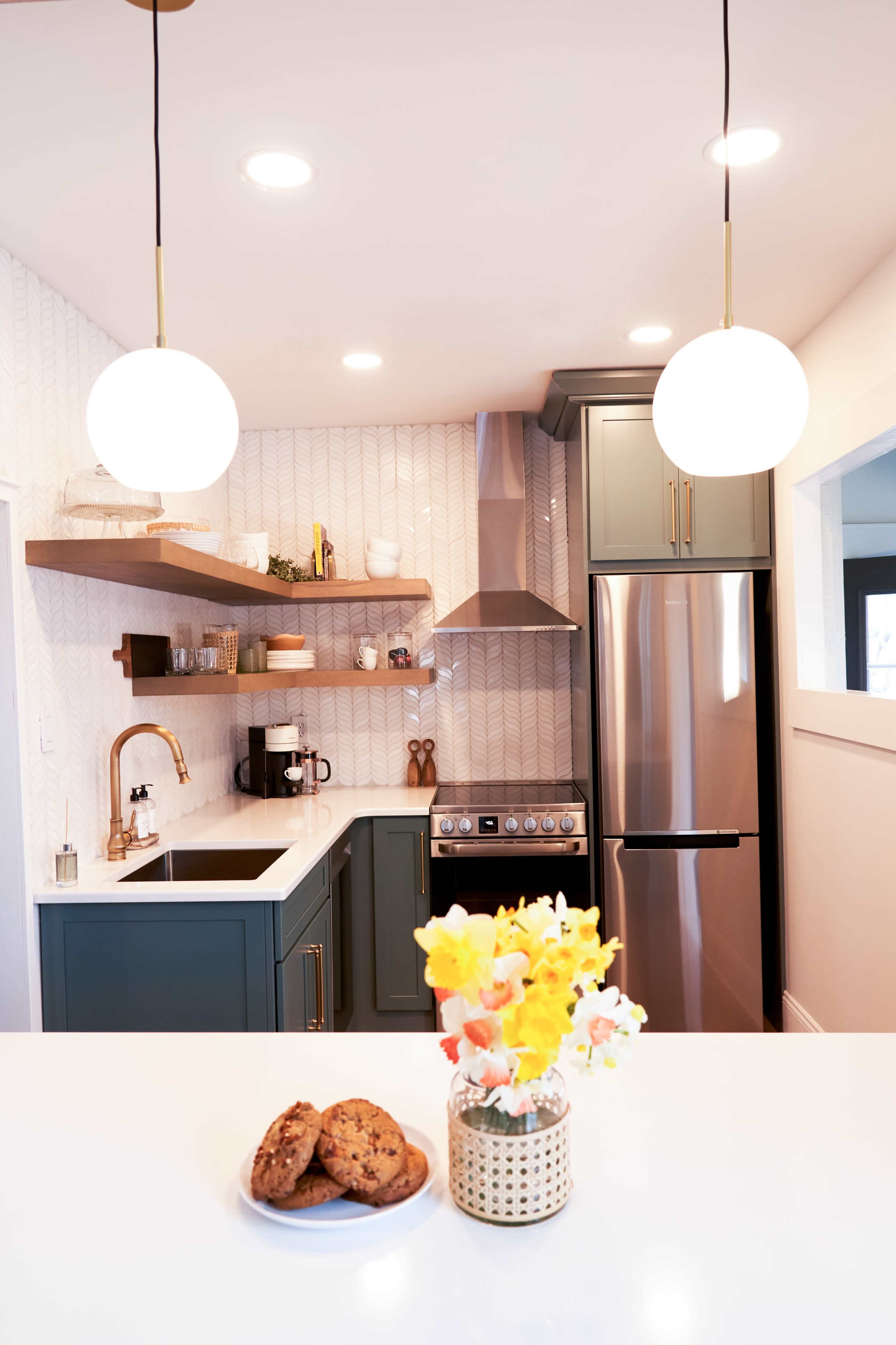 A modern kitchen features green cabinetry, a stainless steel stove, and a countertop with a vase of flowers and a plate of cookies.