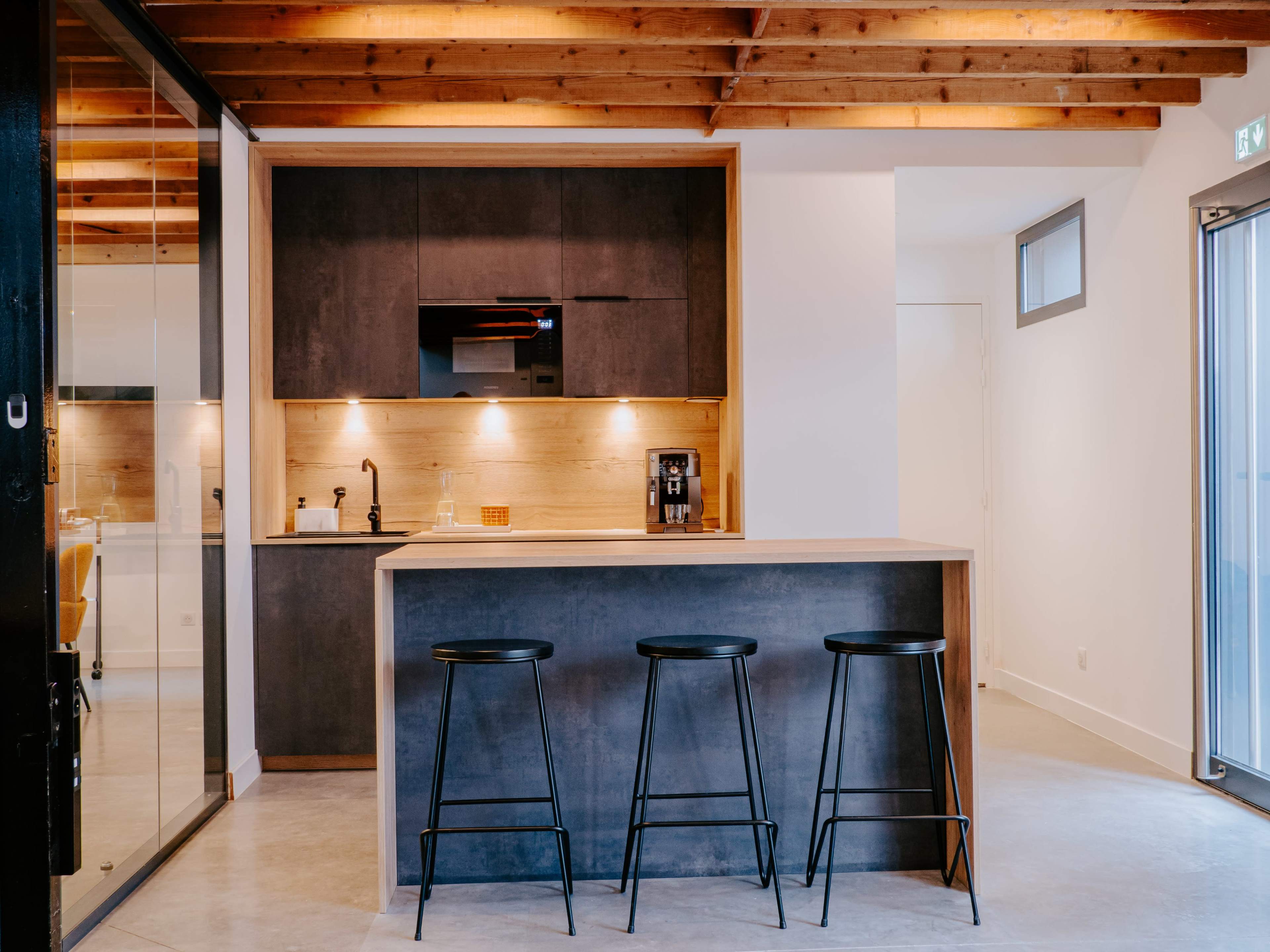 The image shows a modern kitchen with a wooden ceiling, a counter with three black stools, and minimalist cabinetry.
