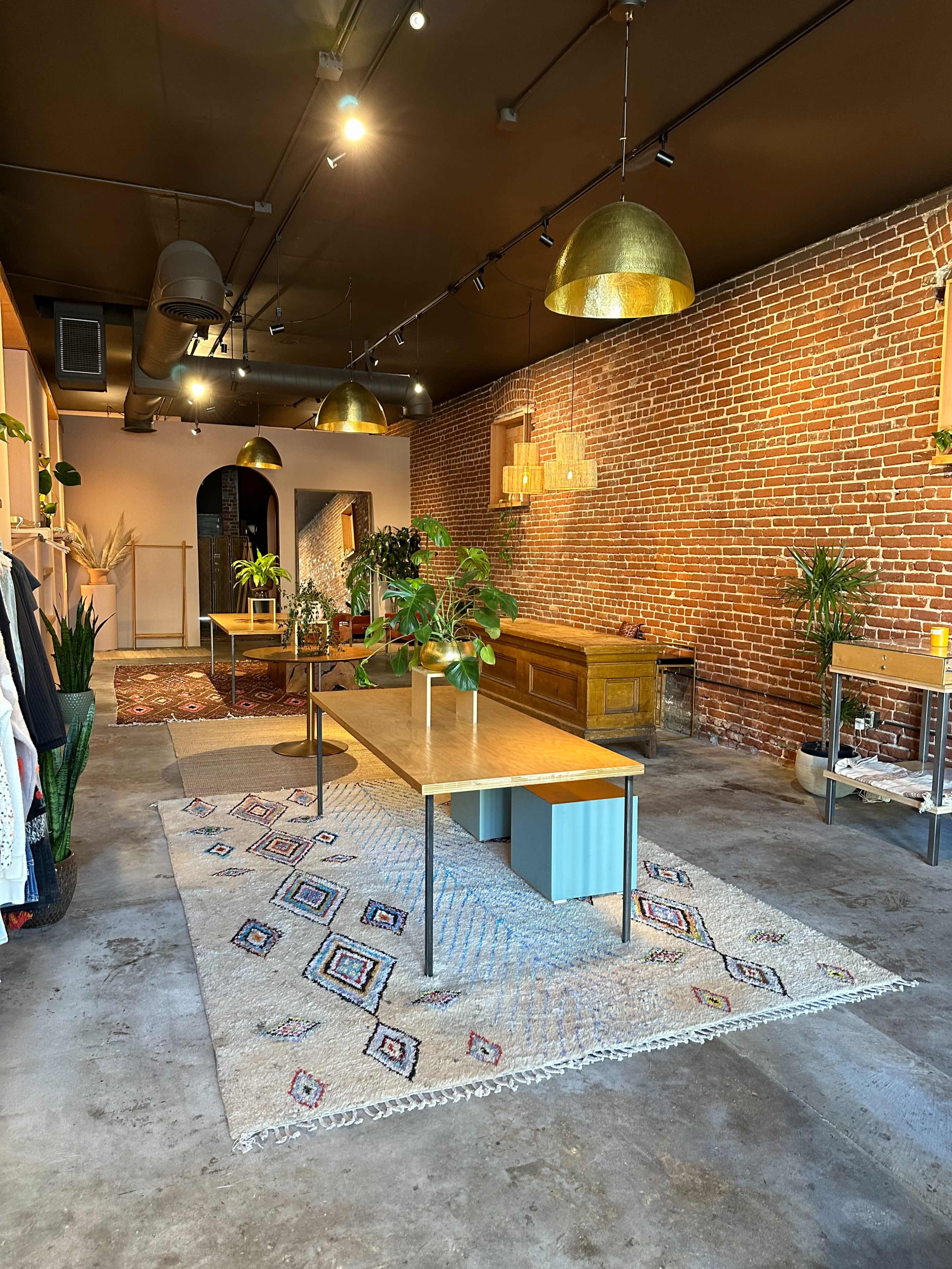 The interior of a shop features exposed brick walls, a large wooden table, and various plants arranged throughout the space.