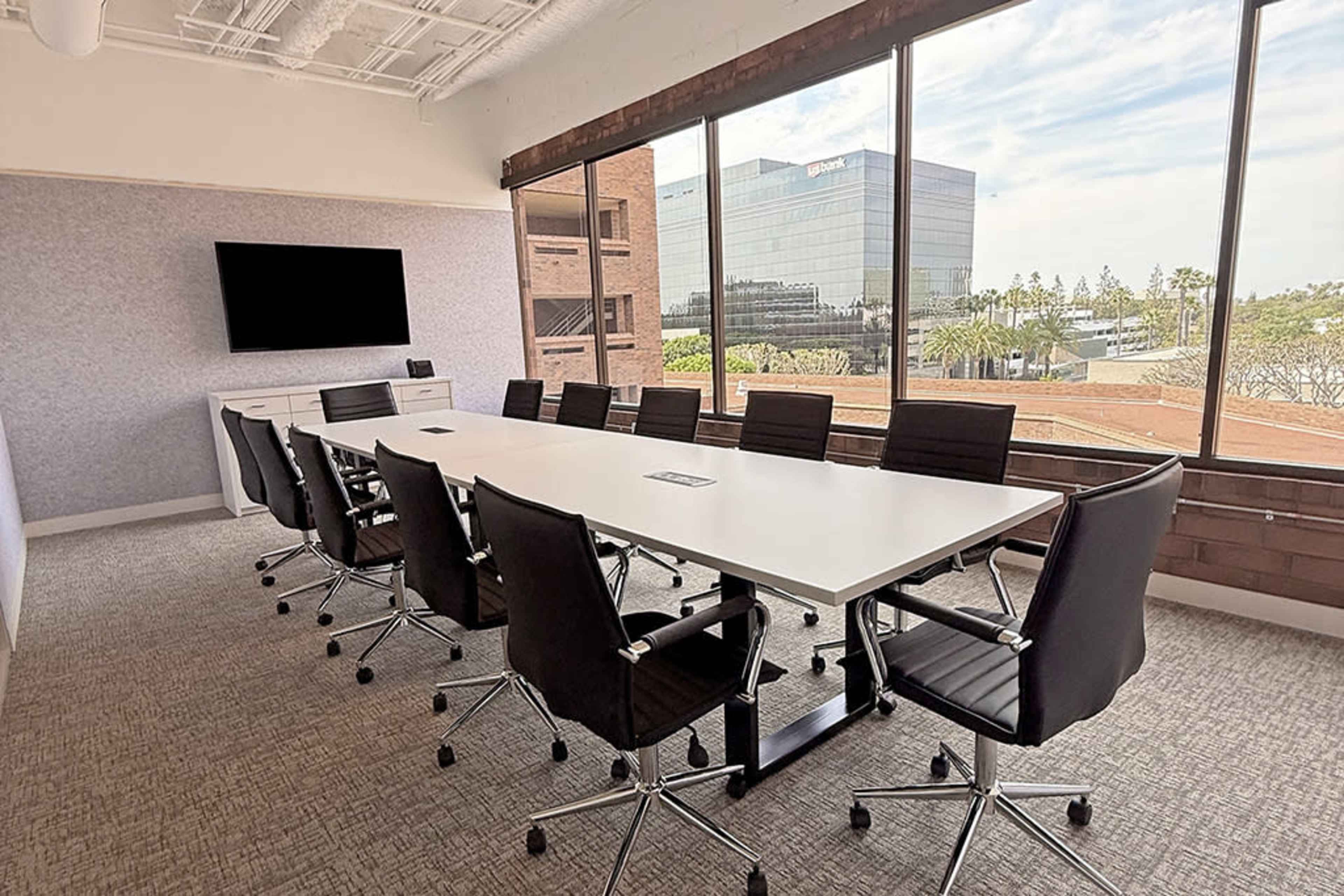 A modern conference room features a large white table surrounded by black swivel chairs, with large windows offering a view of an office building and the outdoors.