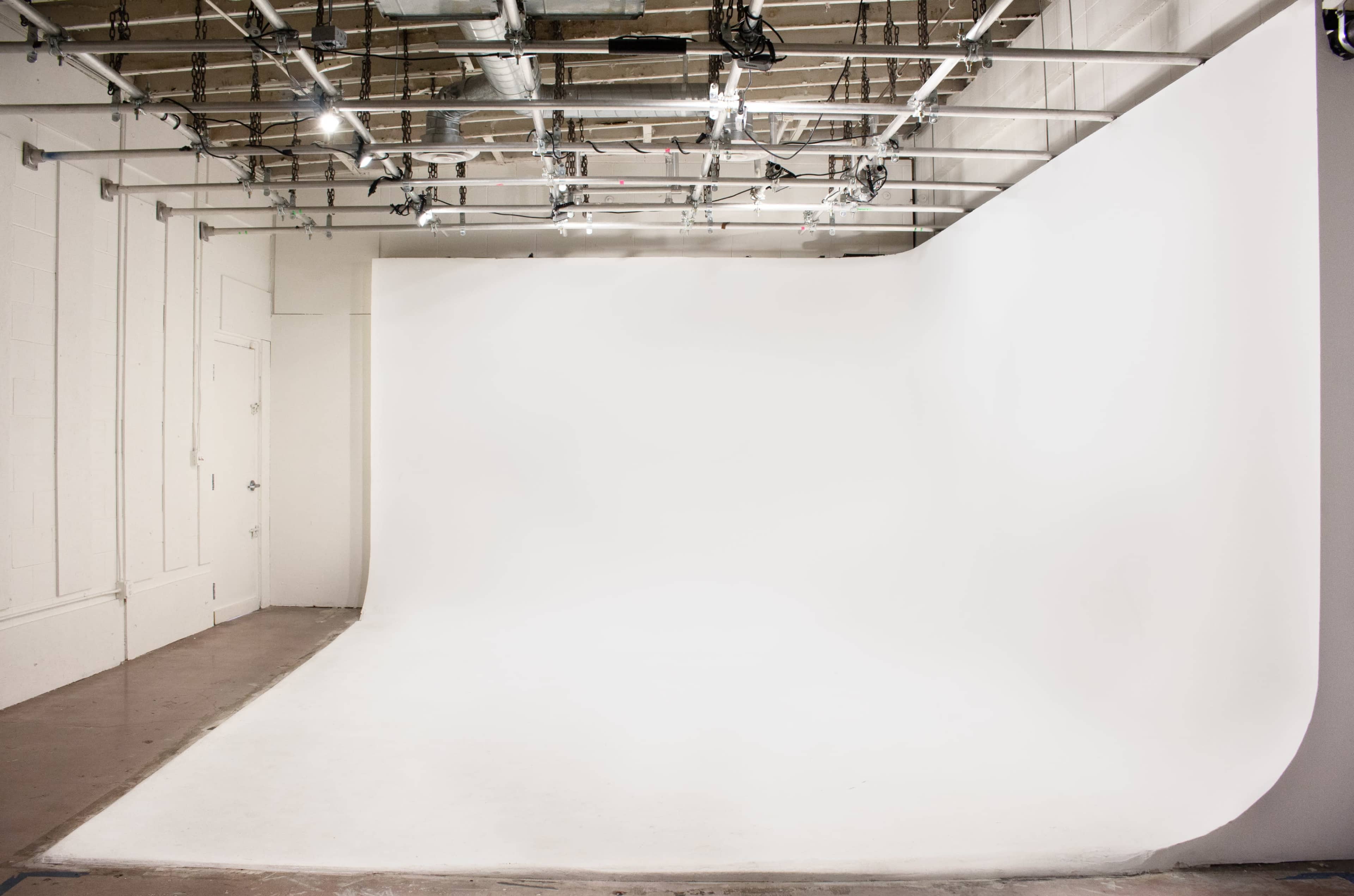 The image shows a spacious photography studio with a seamless white backdrop, polished concrete flooring, and overhead lighting fixtures.
