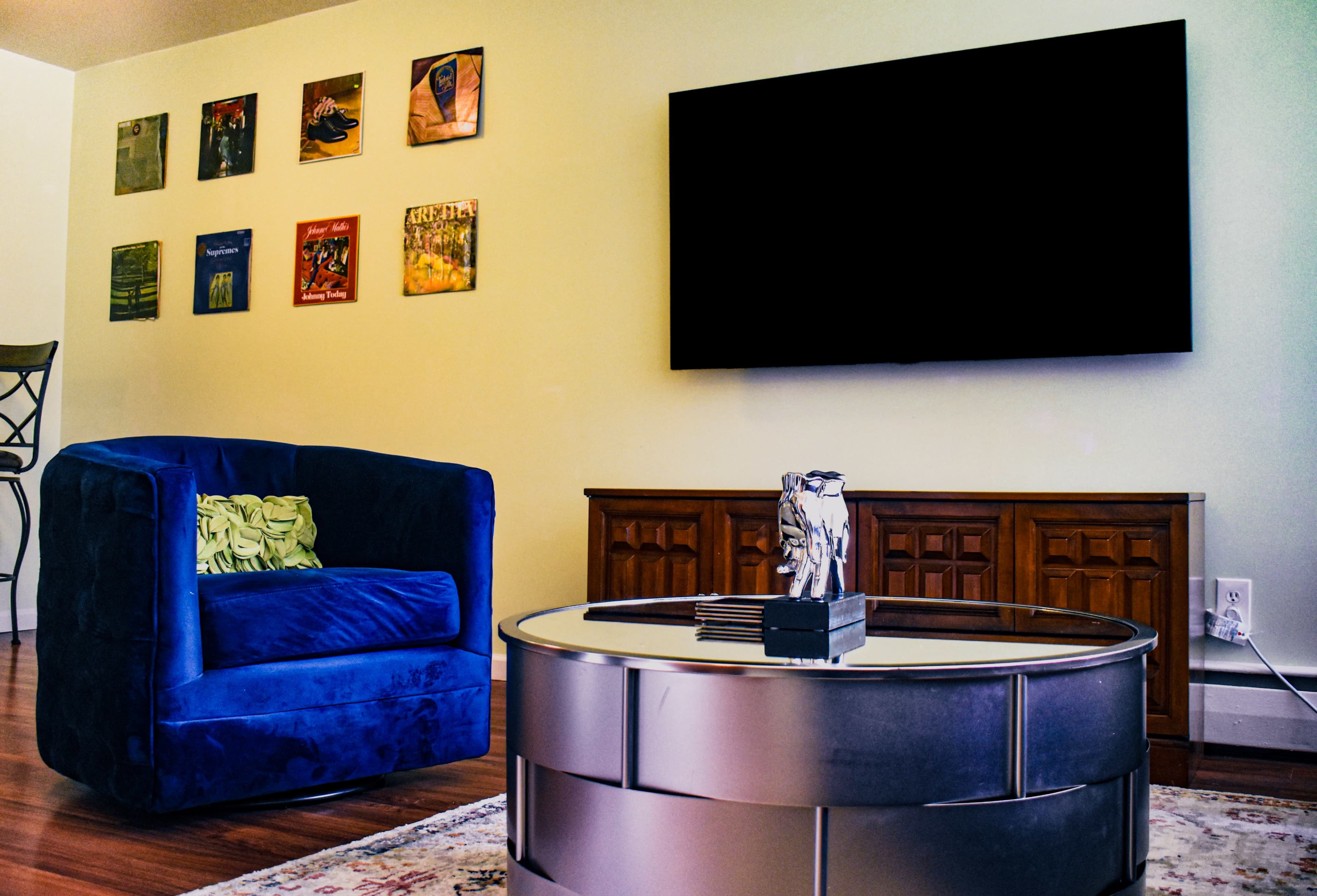 The image shows a living room with a blue armchair, a circular coffee table, and a large flat-screen television mounted on the wall, surrounded by framed art pieces.
