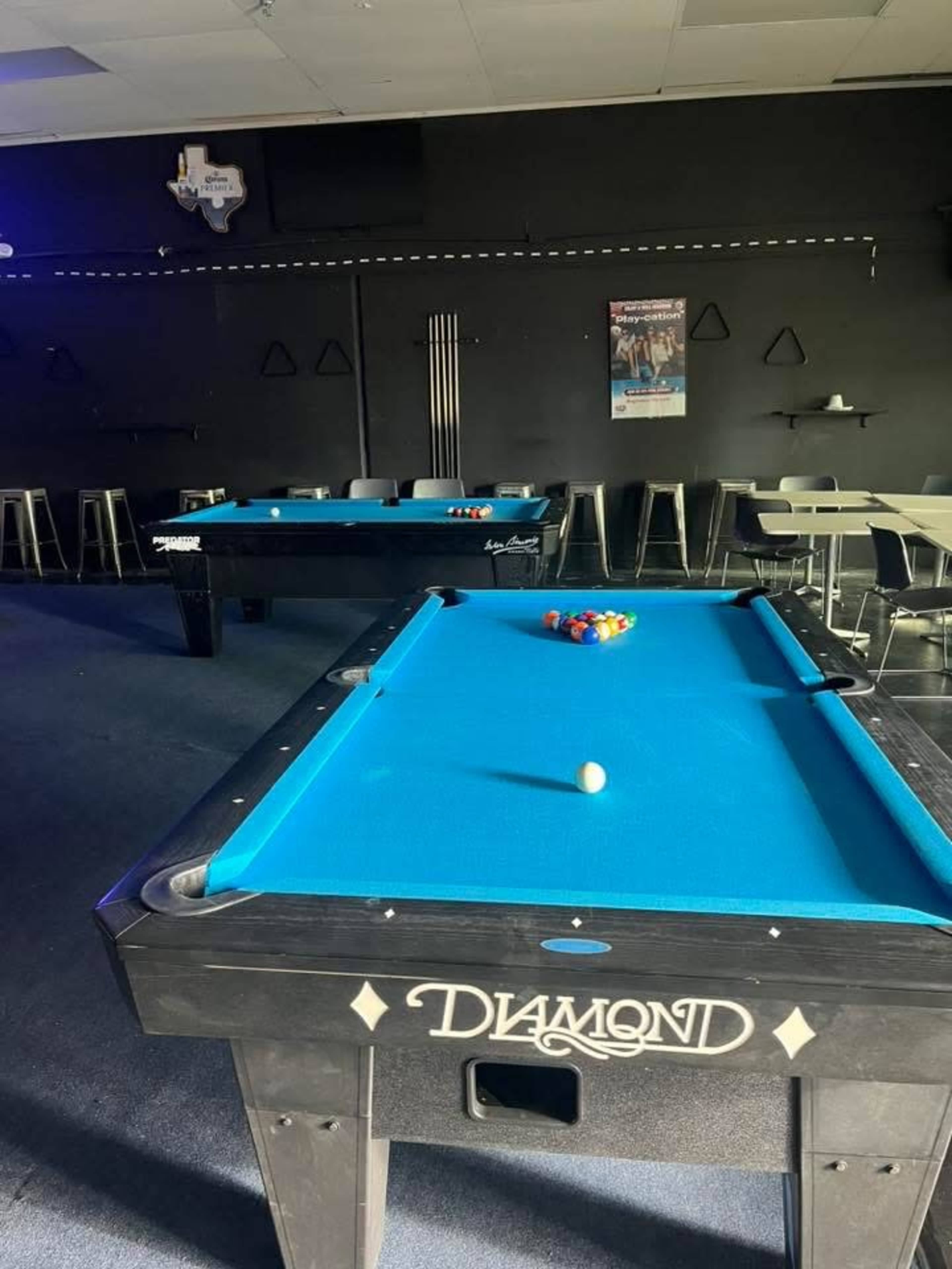 The image shows two pool tables with bright blue cloth in a dimly lit room, with a Texas outline on the wall and a poster in the background.