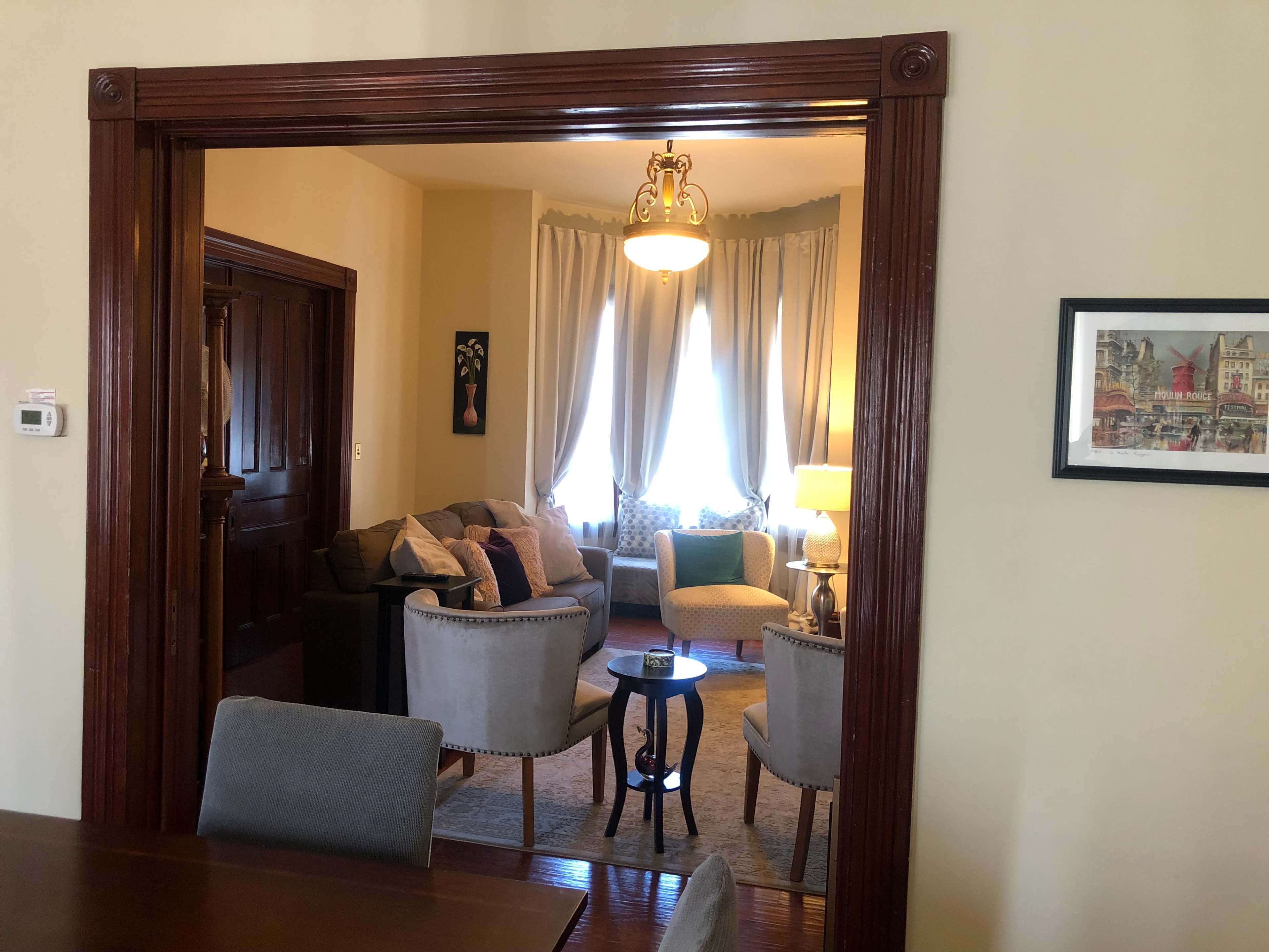 A view through an architectural archway into a living room furnished with a sofa, side chairs, and a lamp, illuminated by natural light from large windows.