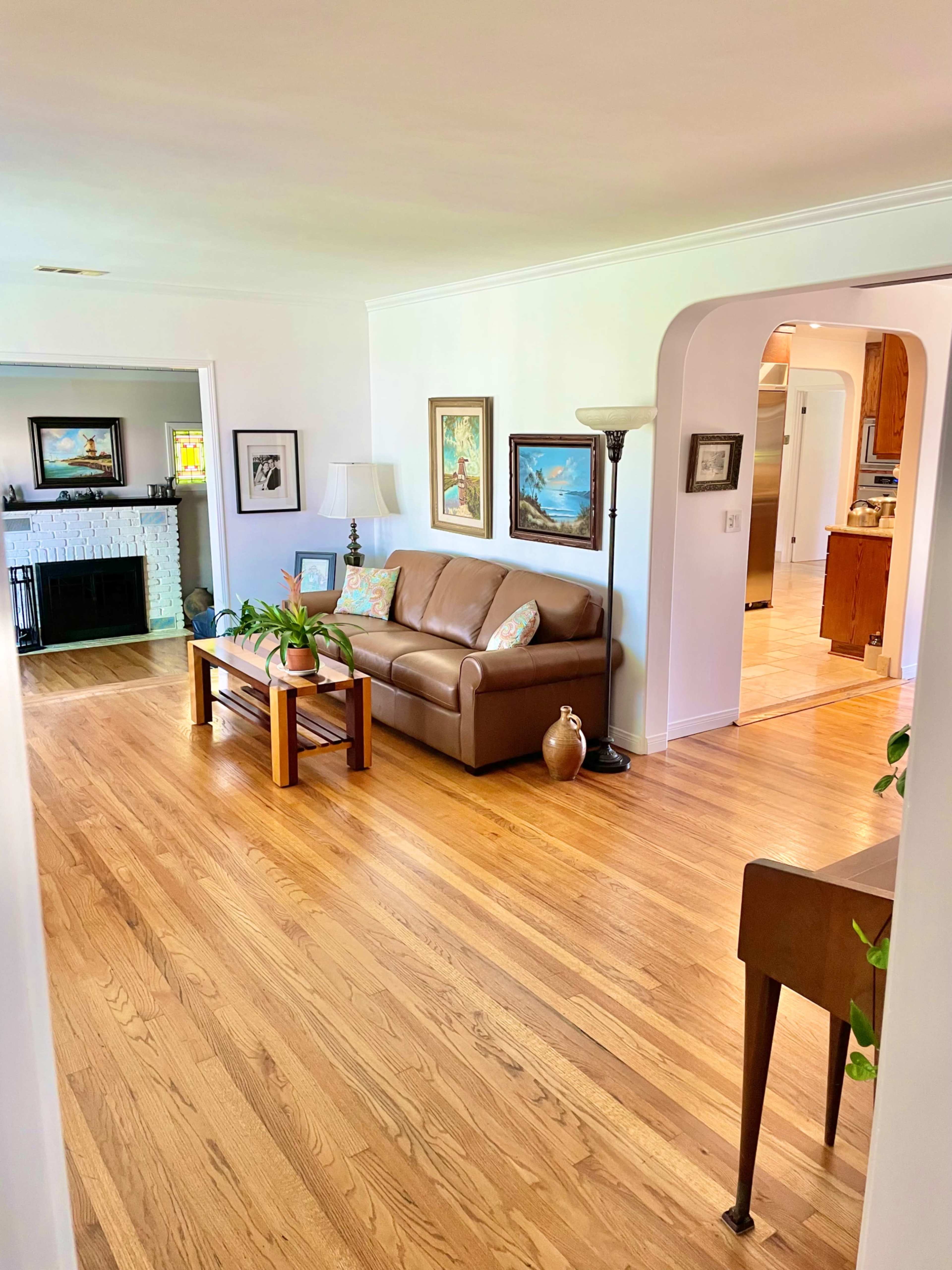 A living room with a brown sofa, a wooden coffee table, and framed artwork on the walls, leading into a kitchen area.