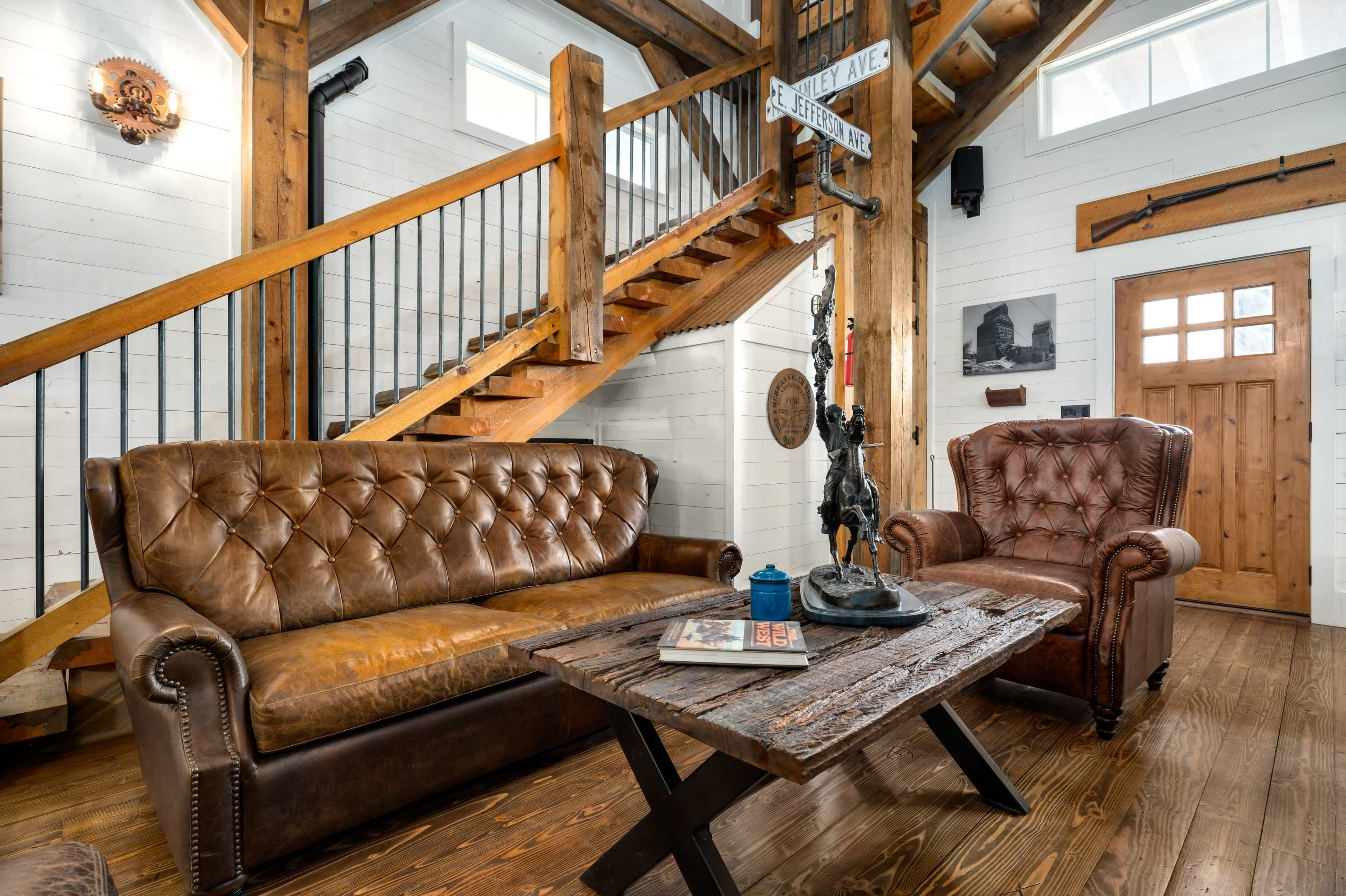 A rustic living area with a leather sofa and armchair, a wooden table, and a staircase leading to an upper level.