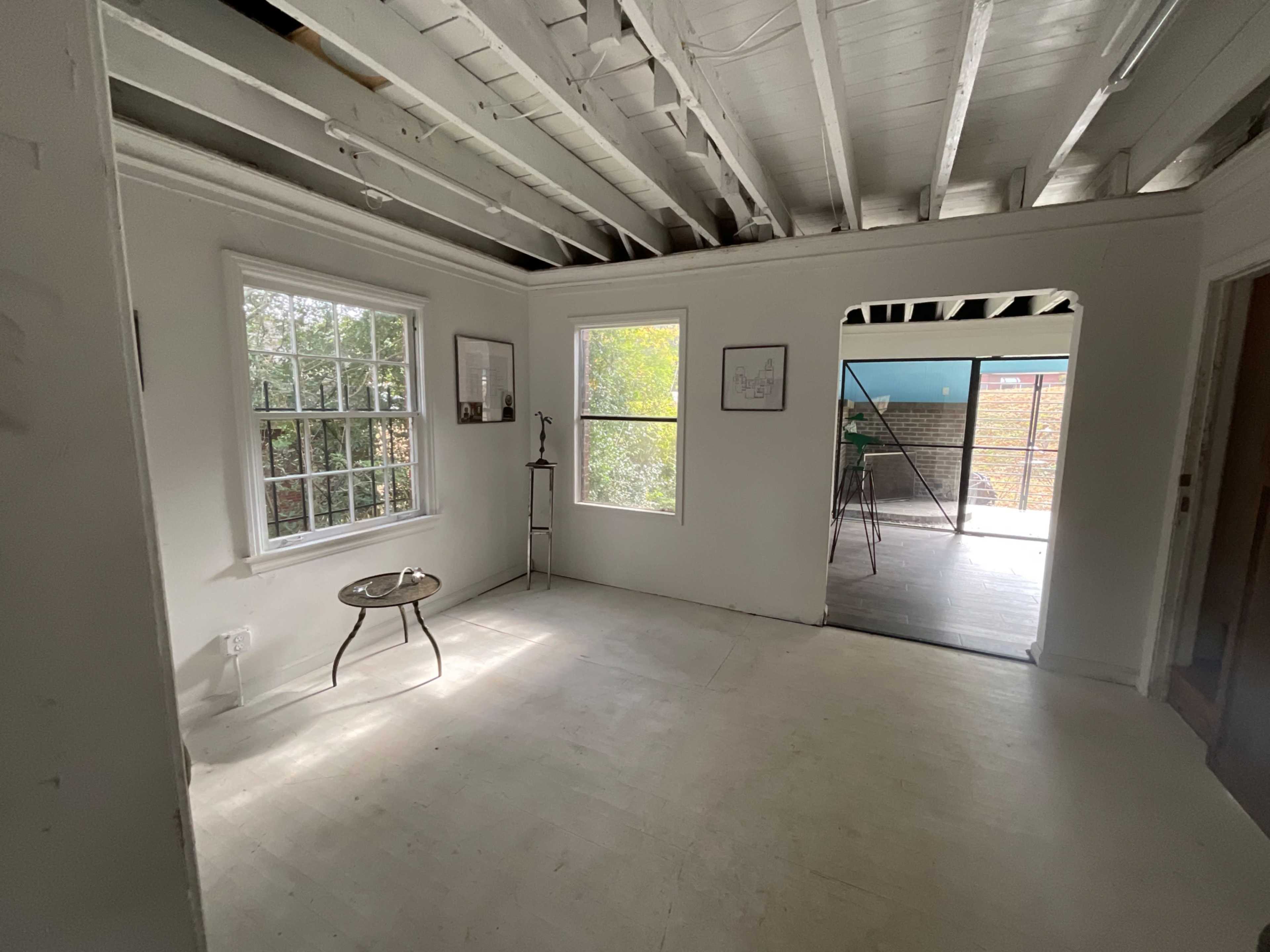 The image shows a bright, empty room with white walls, exposed beams, and a small round table near a window.