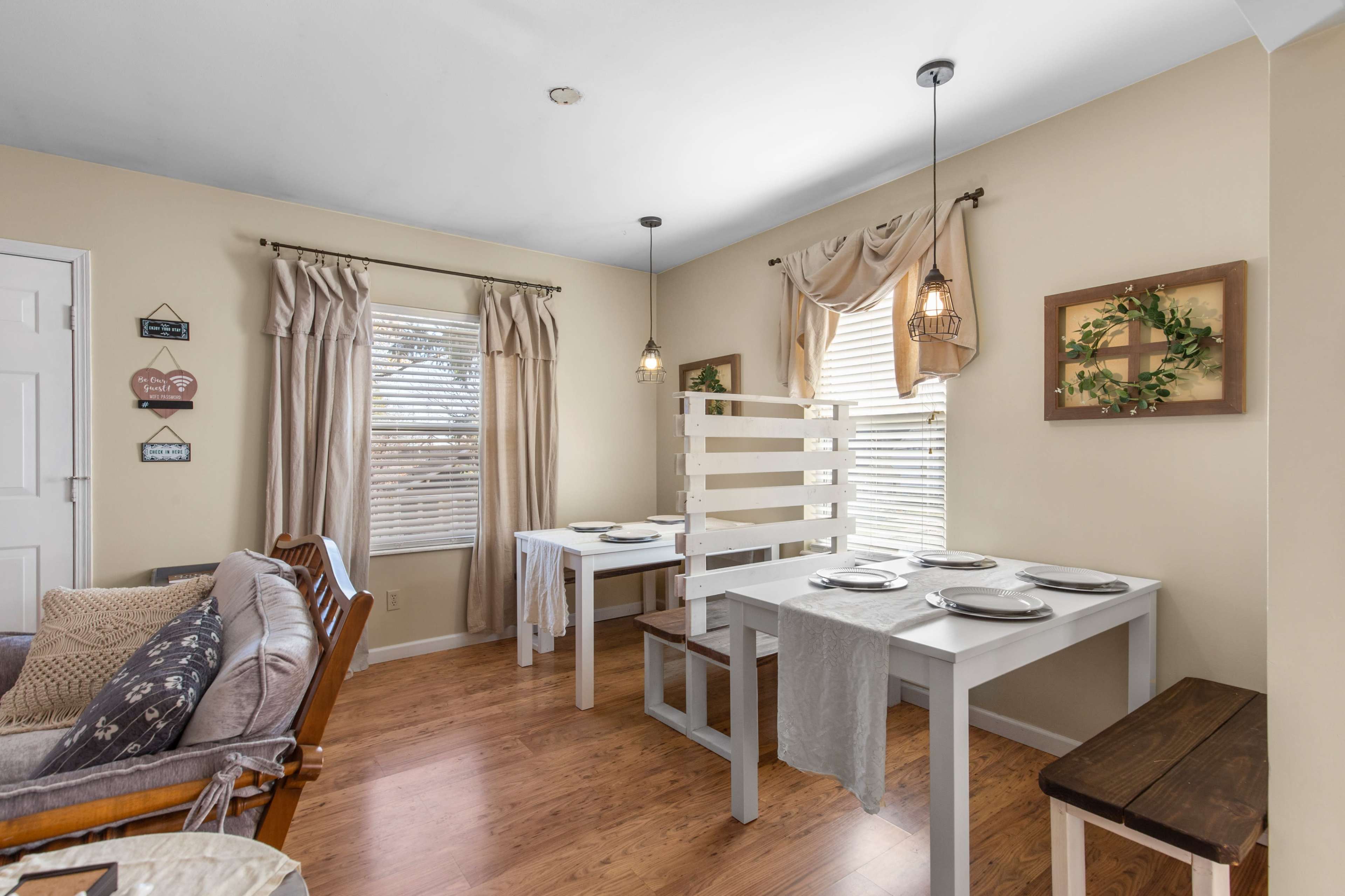 A cozy dining area features two white tables with set plates, surrounded by beige walls and warm wood flooring.