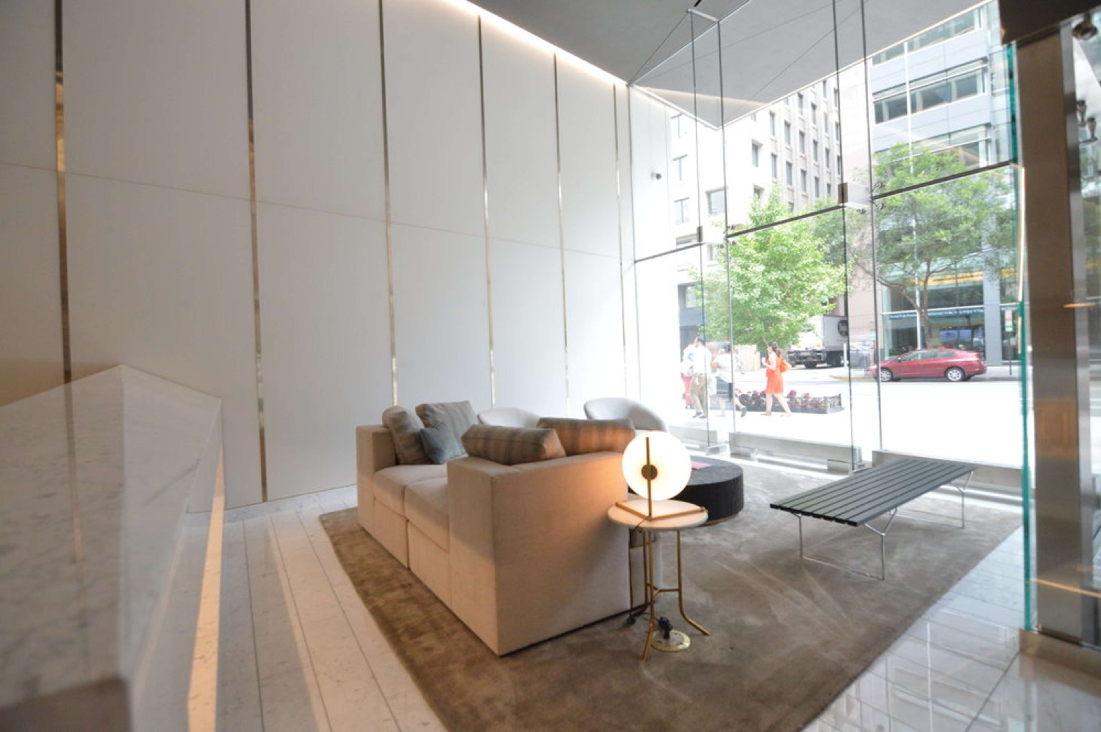 A modern, minimalist living area with a beige sofa, a round coffee table, and large glass windows overlooking a city street.