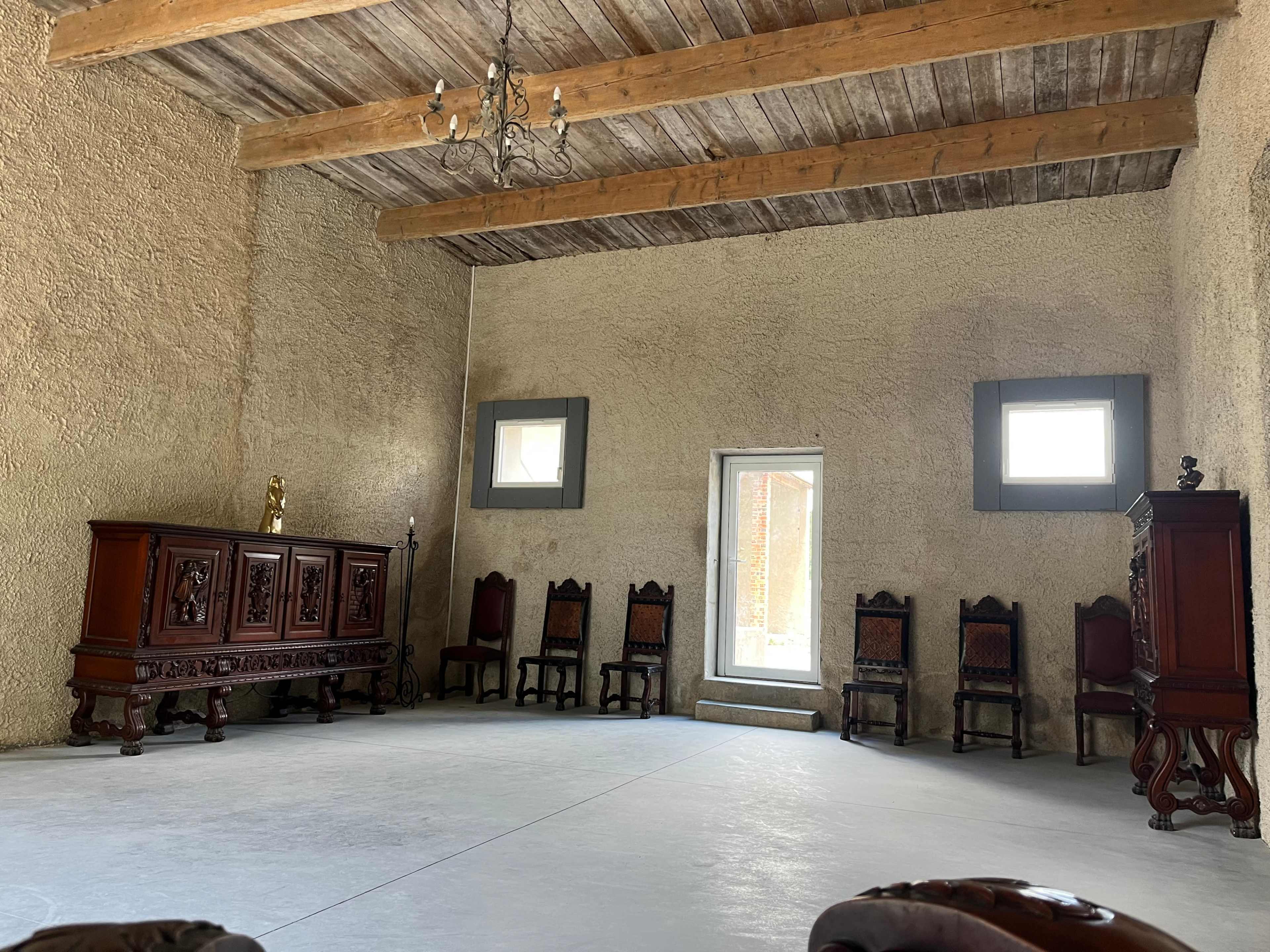 The image shows a spacious room with a high wooden-beamed ceiling, featuring a large wooden sideboard, several chairs, and a window letting in natural light.