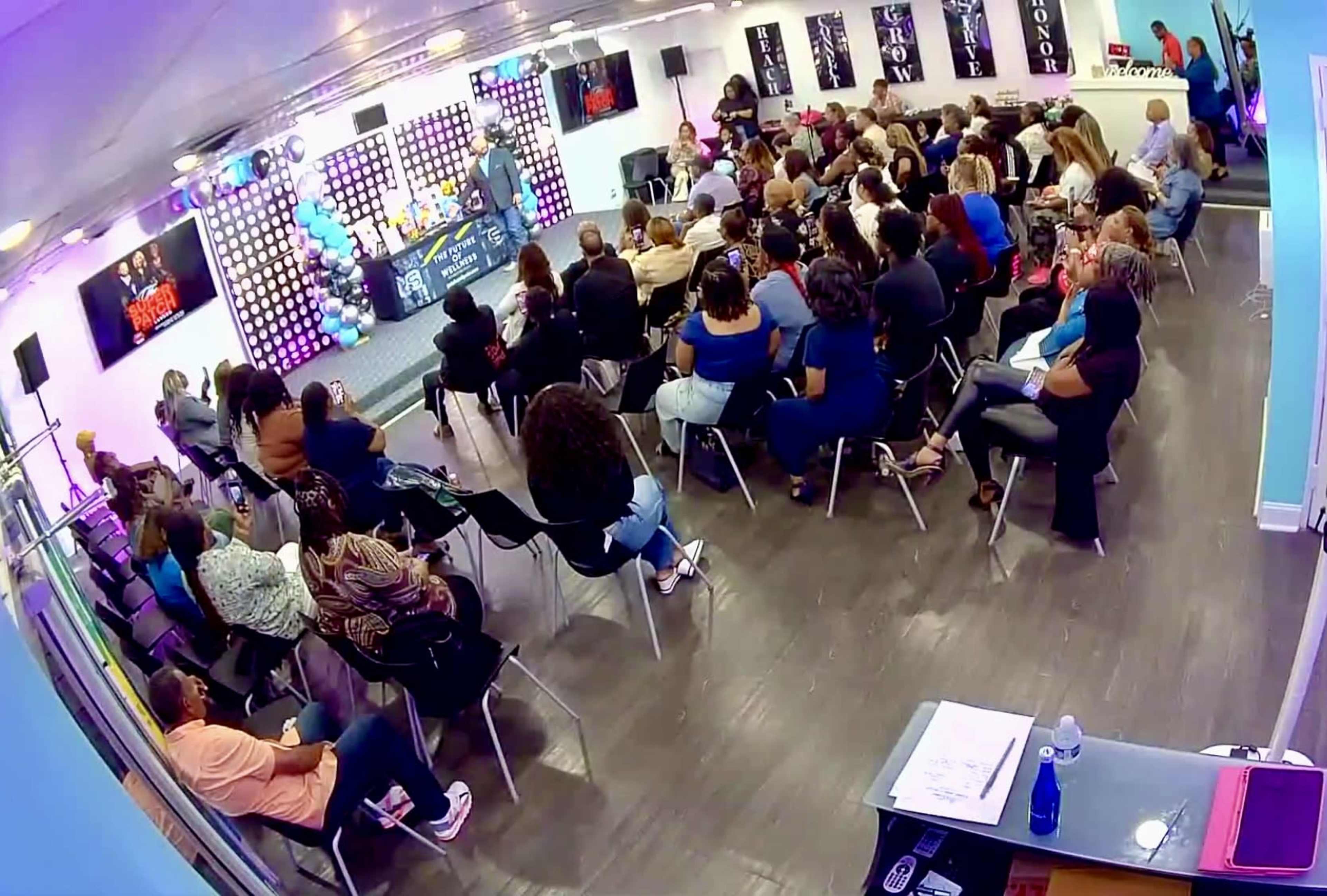A large gathering of people is seated in a room facing a brightly decorated stage with colorful lights and a speaker addressing the audience.