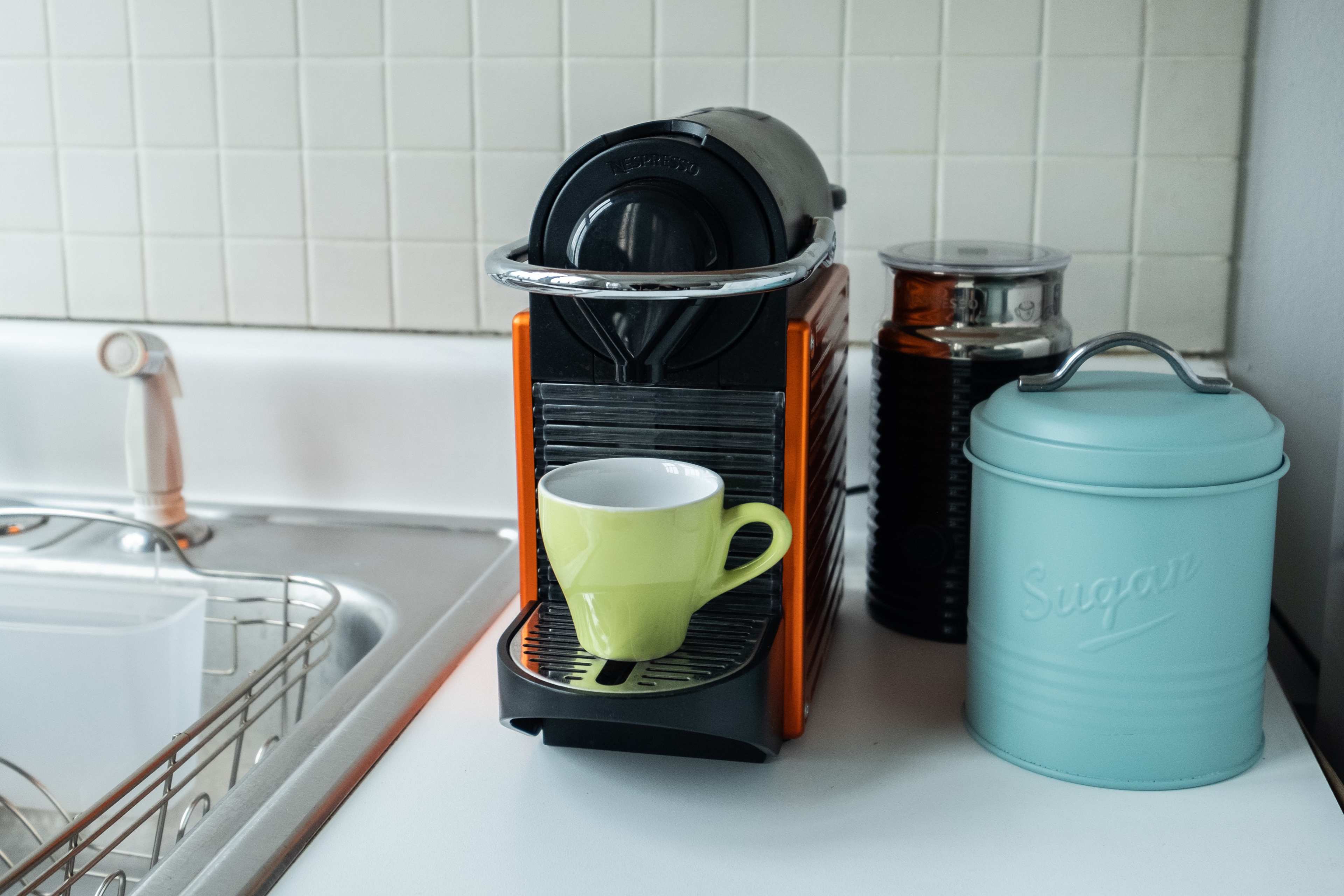 The image shows a kitchen counter with an orange coffee machine next to a green cup, a sugar canister, and a jar.
