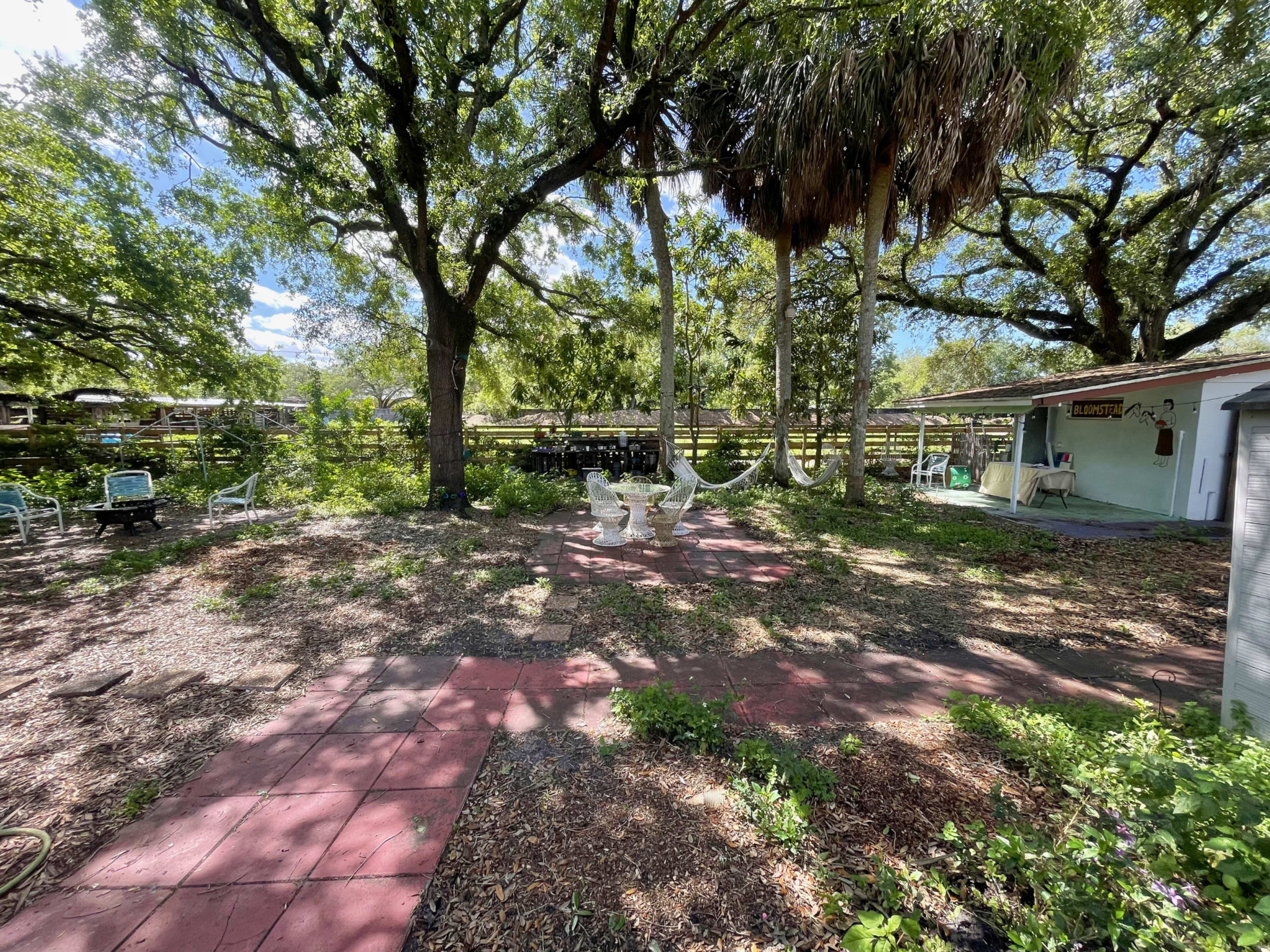 The image shows a grassy outdoor area with a circular arrangement of white chairs surrounded by trees and a small building in the background.