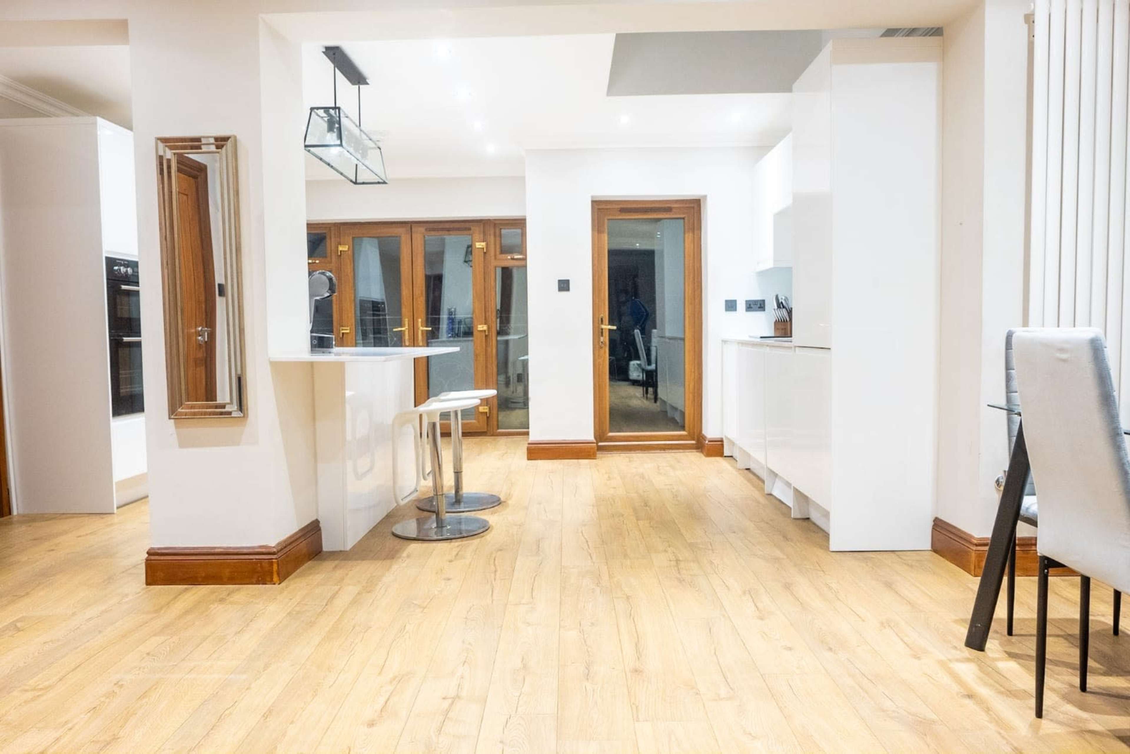 The image shows a modern kitchen area with wooden flooring, white cabinetry, and glass doors leading to an outdoor space.