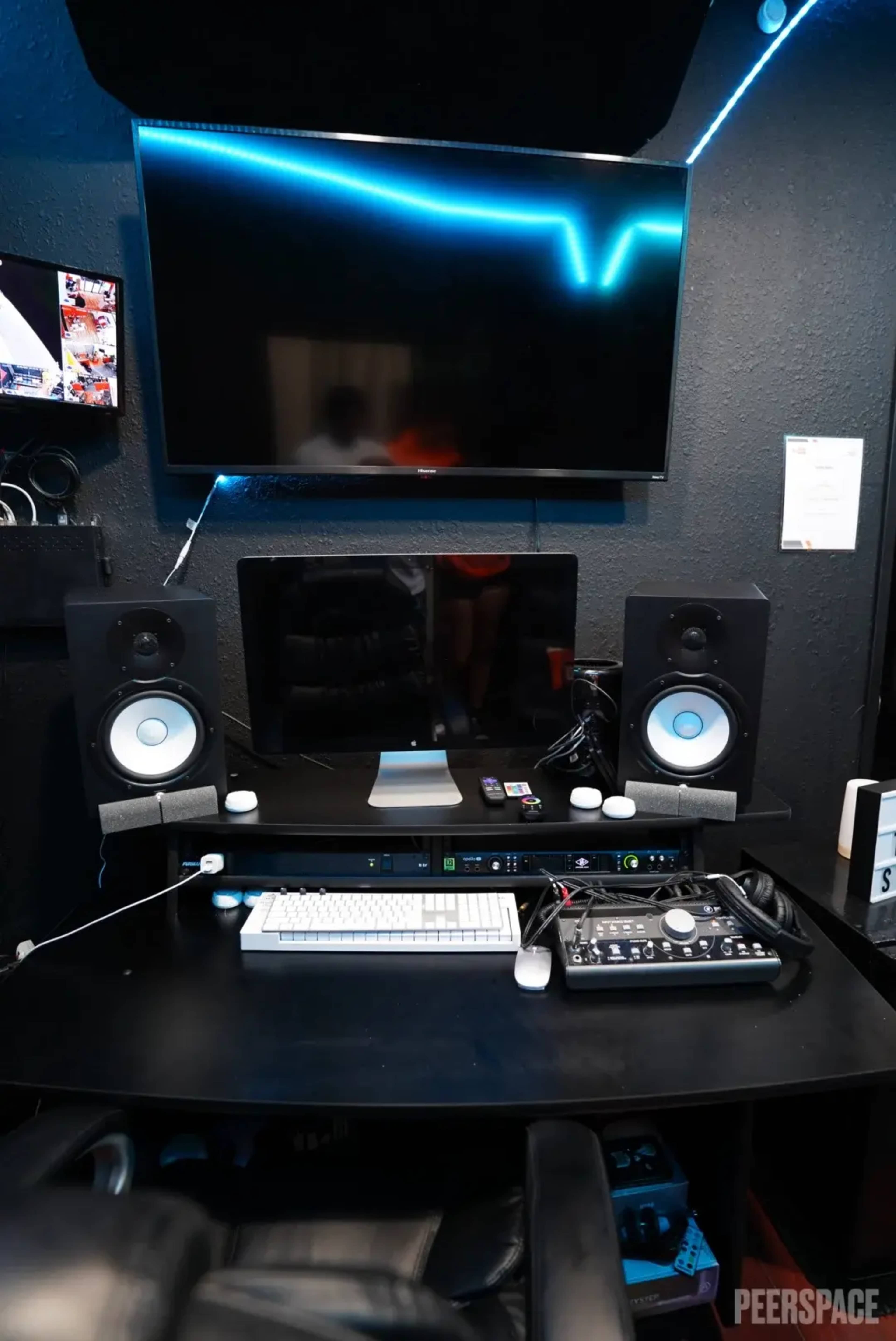A music production studio setup features a black desk with speakers, a computer, a mixing console, and a large television mounted on the wall.