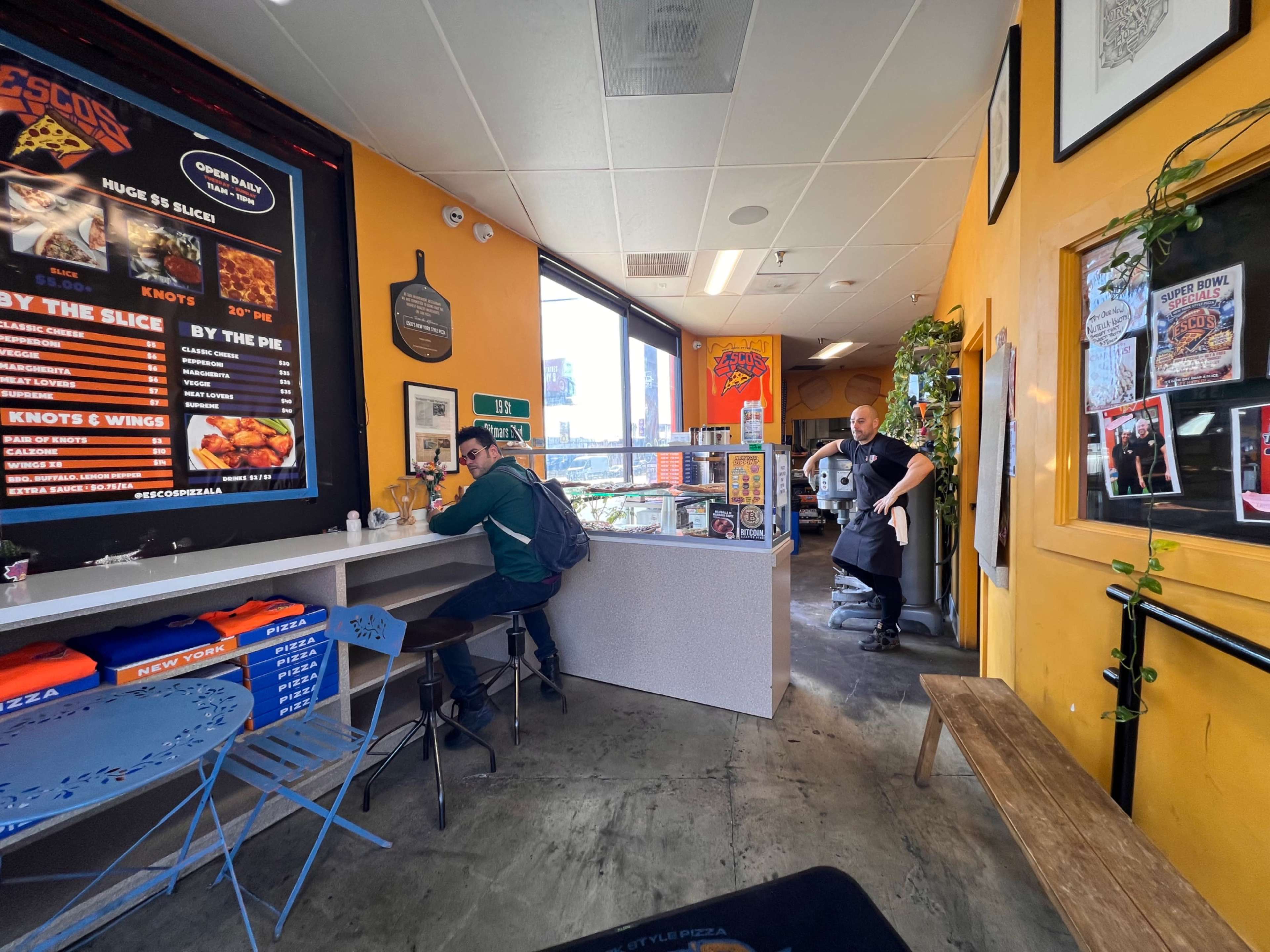 The image shows an interior of a casual dining space with a bright yellow wall, featuring a man walking past a customer seated at a counter, while various food menus are displayed on the walls.