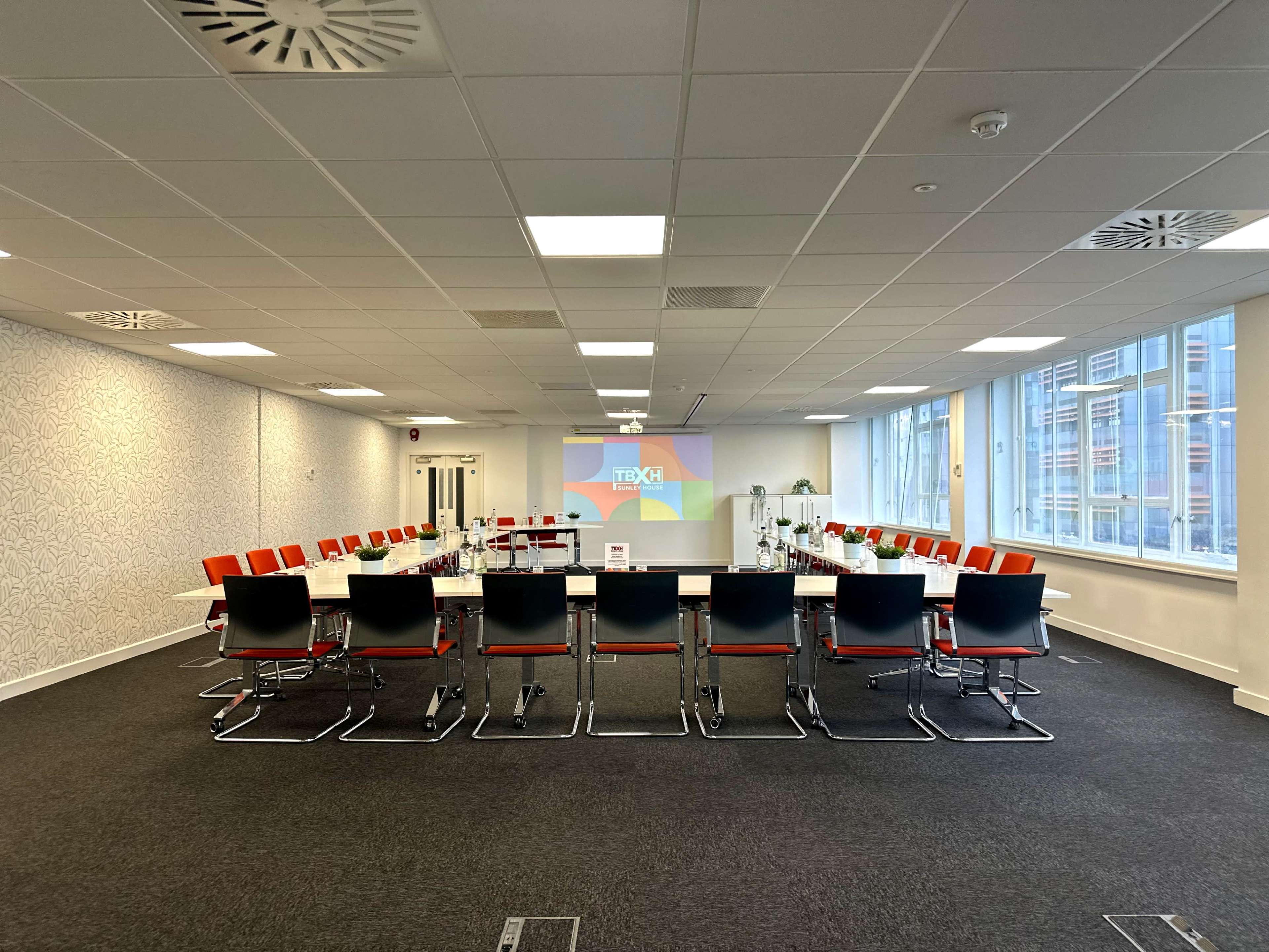 A modern conference room is set up with a large rectangular table surrounded by black and red chairs, with a projector screen displaying a colorful presentation in the background.