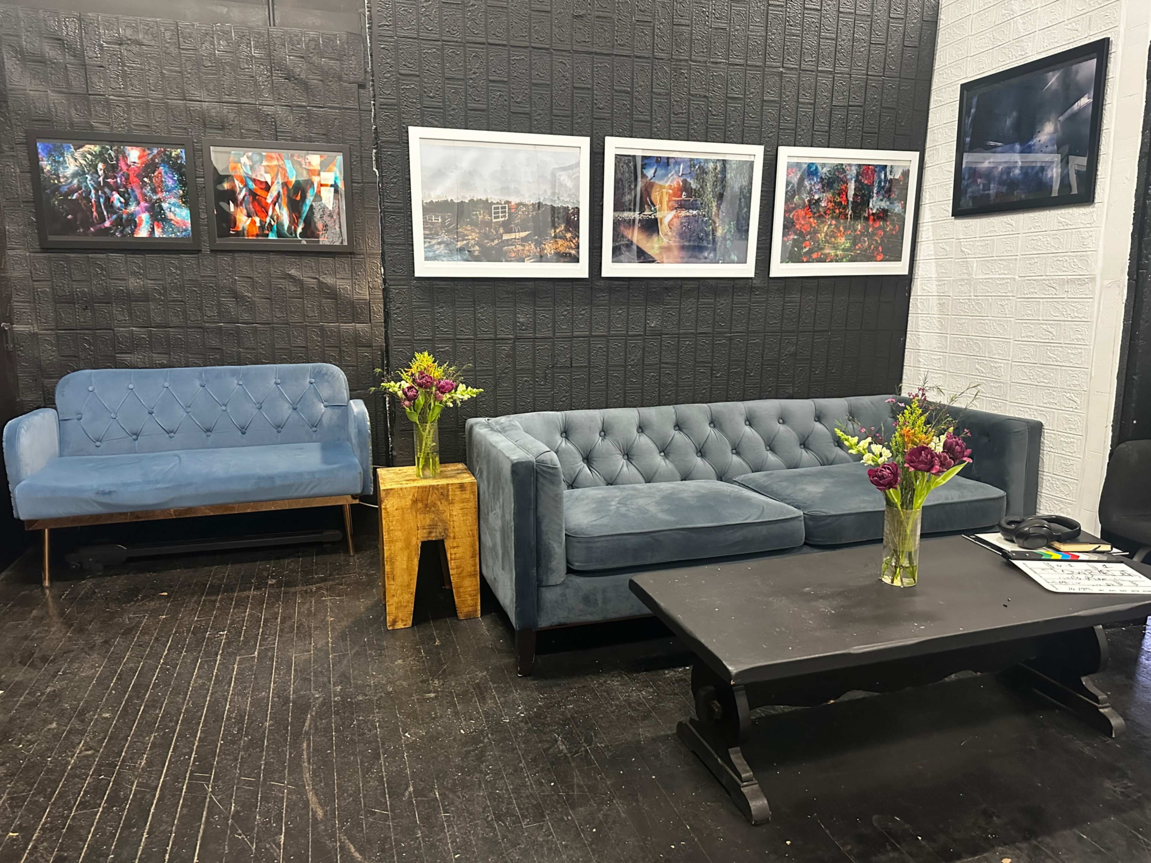 The scene features a stylish seating area with a blue tufted sofa, a matching loveseat, a wooden side table with a vase of flowers, and several framed photographs hanging on the wall.