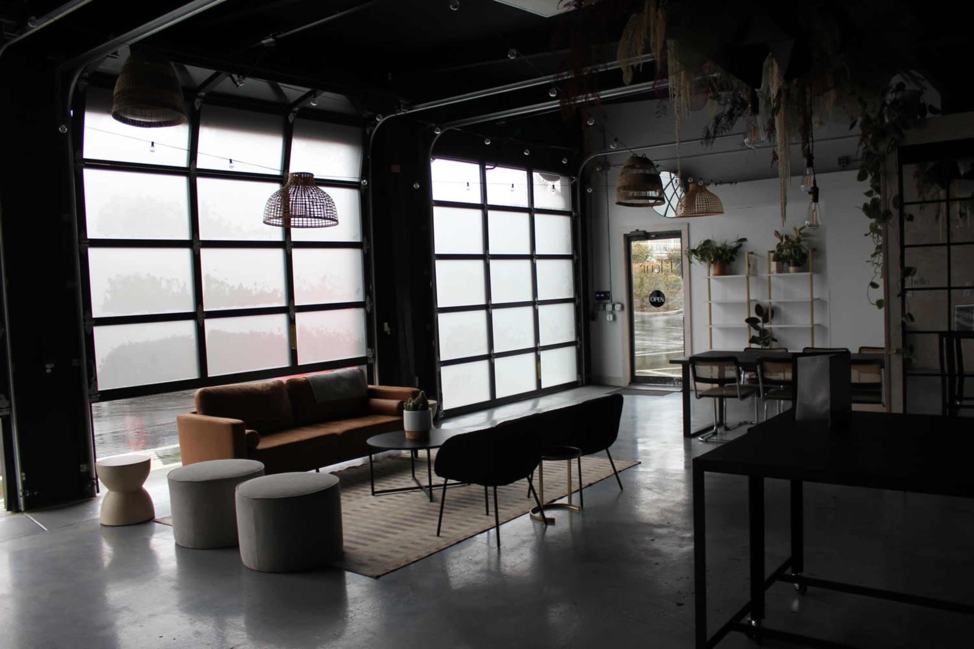 The image shows a modern interior space with large windows, a brown sofa, black chairs, and a minimalist decor featuring plants and tables.