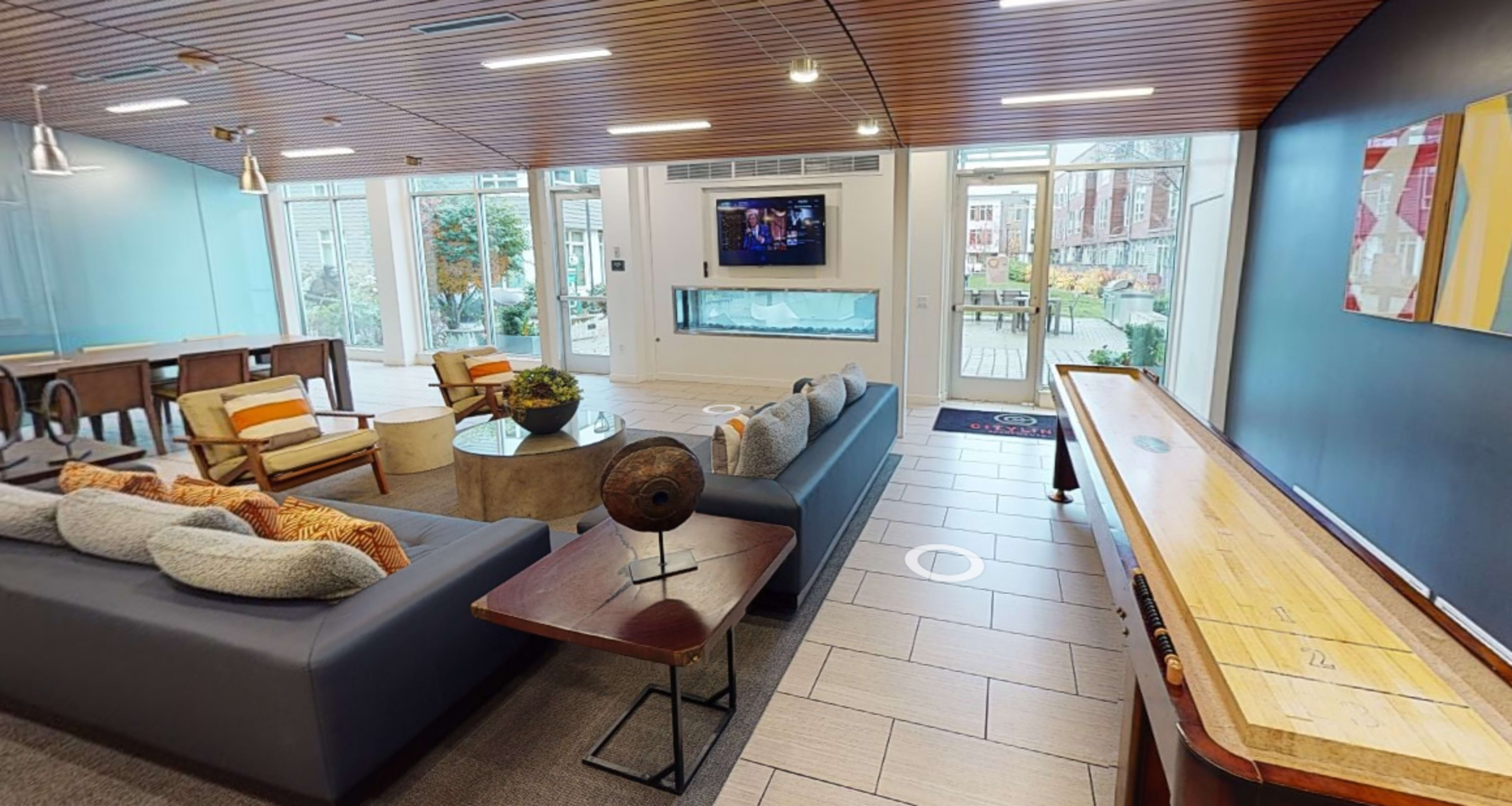 A modern lounge area with a sofa, a coffee table, a television, and a shuffleboard table, all set against large windows revealing an outdoor view.