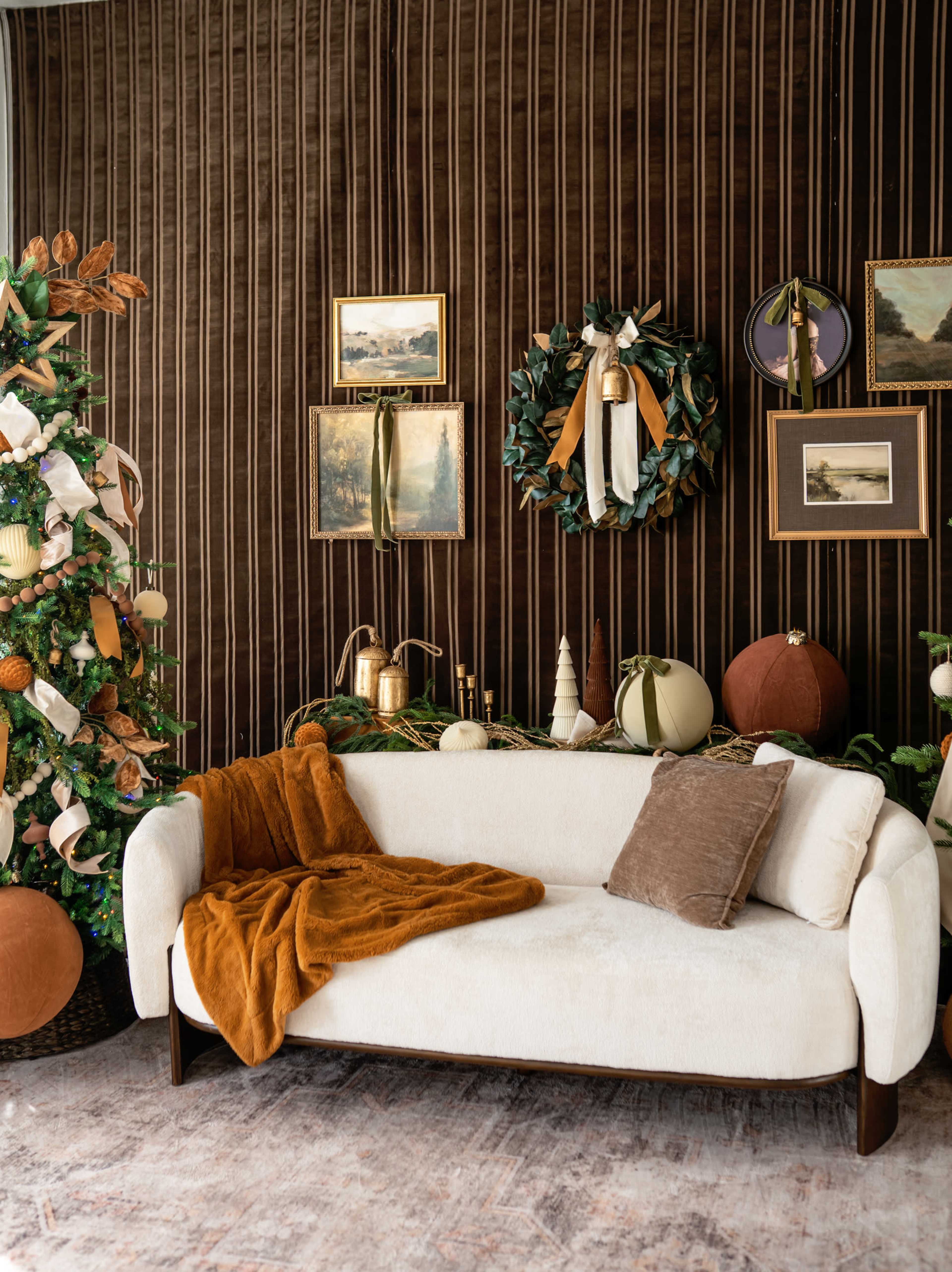 A cozy living room features a white couch with a brown throw blanket, in front of a striped dark wall adorned with framed artworks, a decorative wreath, and festive seasonal decor.