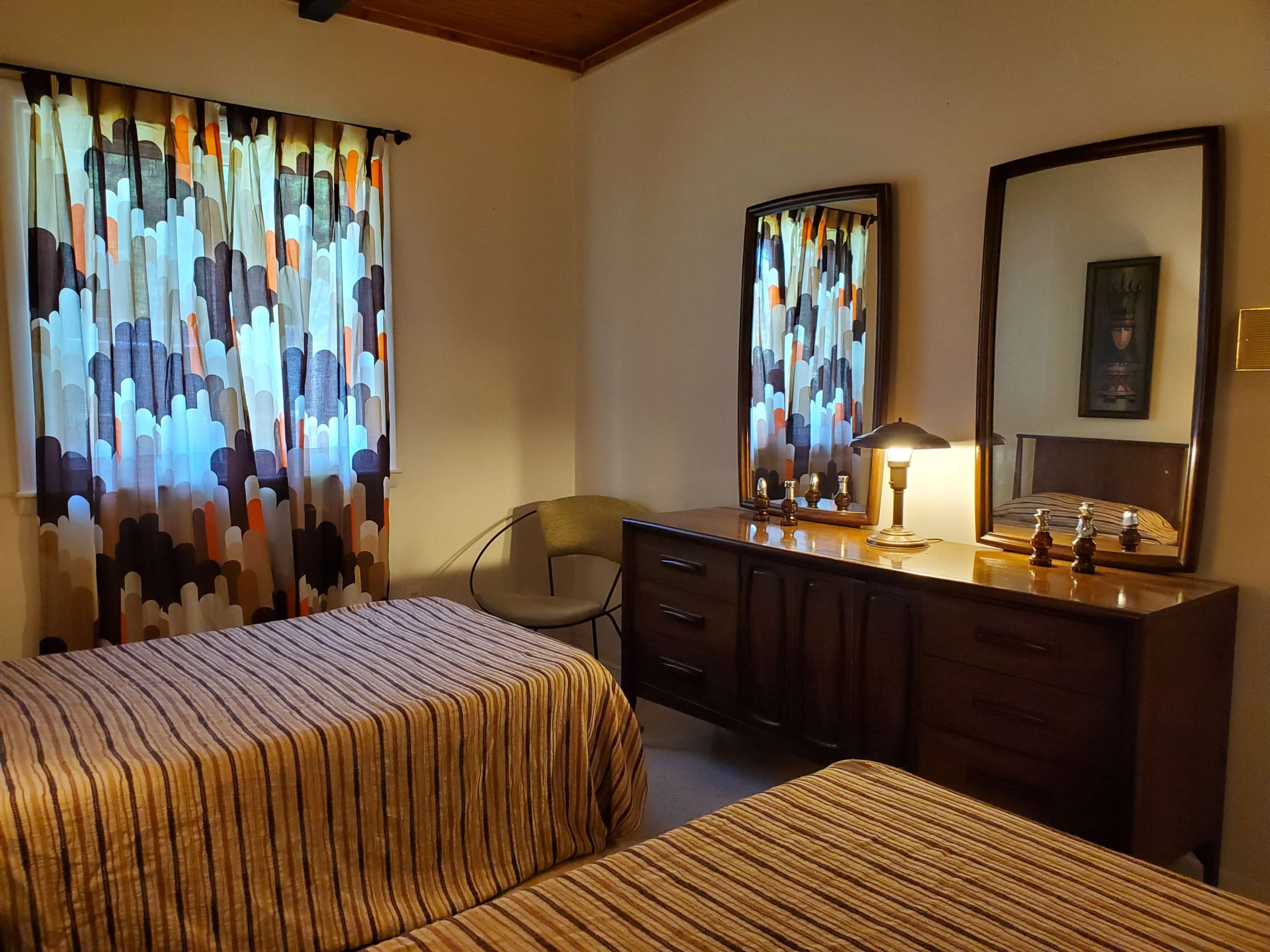 A bedroom with two striped beds, a wooden dresser, two mirrors, and a lamp on a bedside table.
