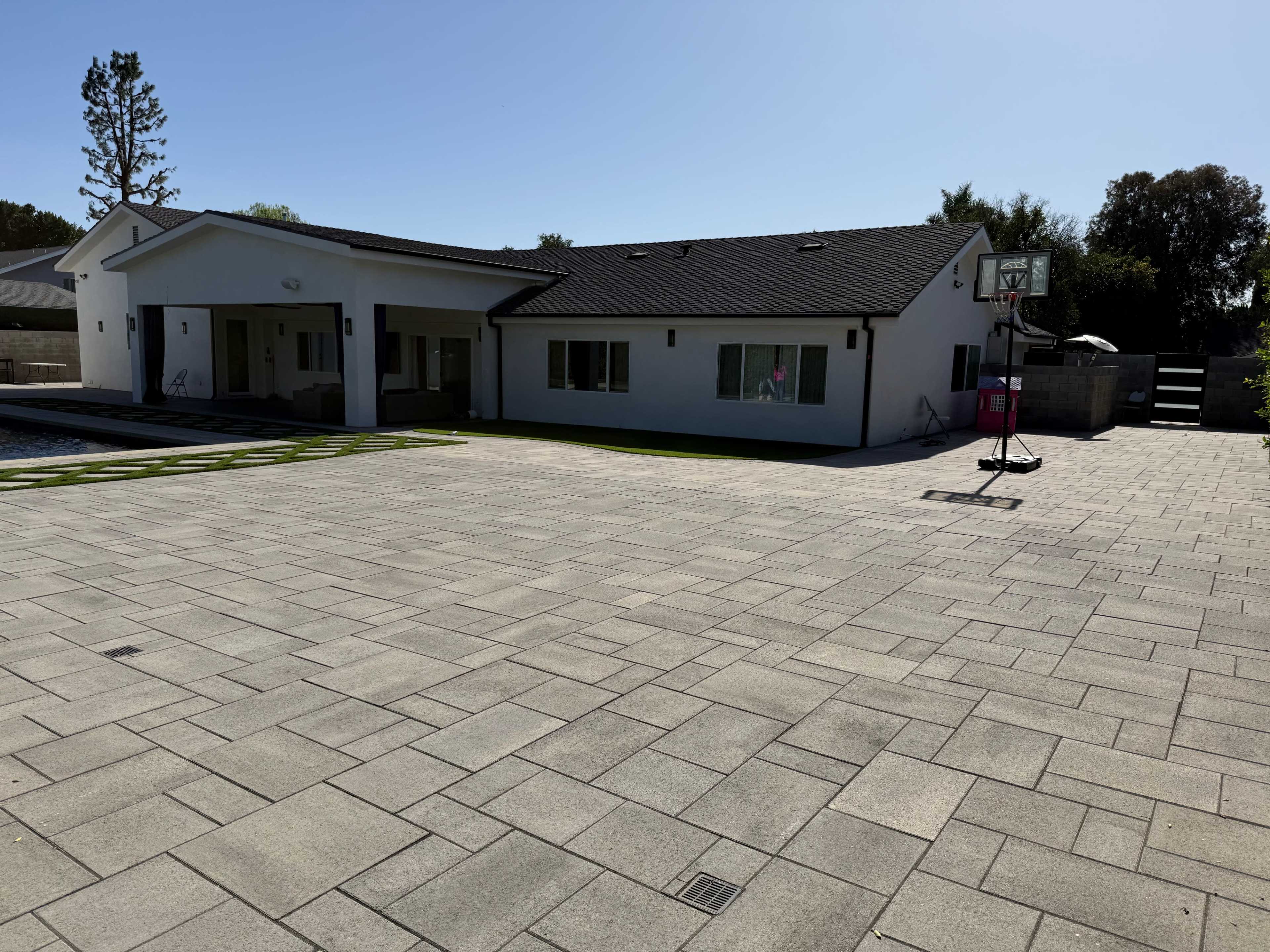 The image shows a modern single-story house with a basketball hoop in the driveway and an expansive paved area in front.