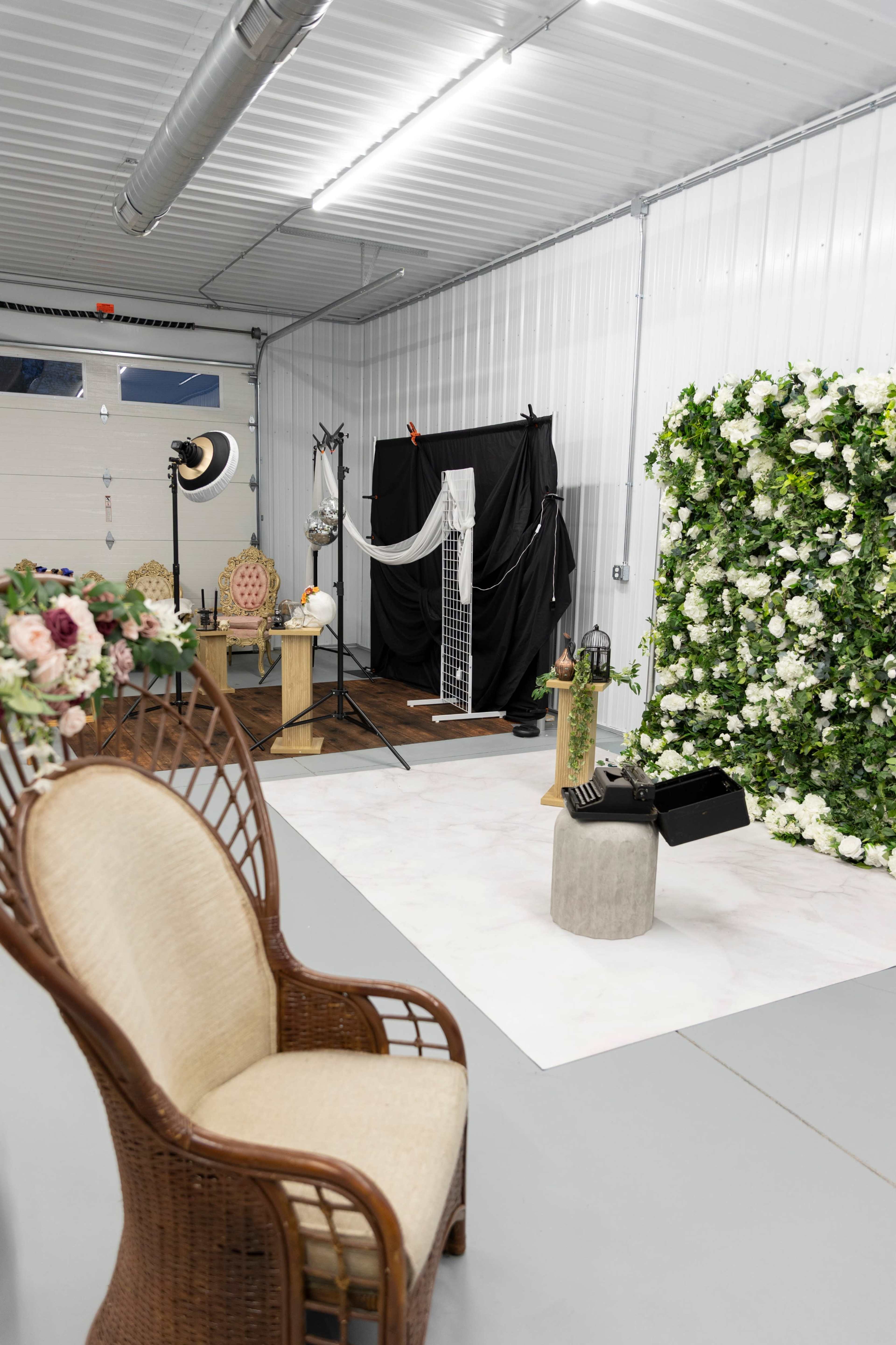 The image shows a photography studio setup featuring a green floral wall, a white backdrop, various props, and seating arrangements.