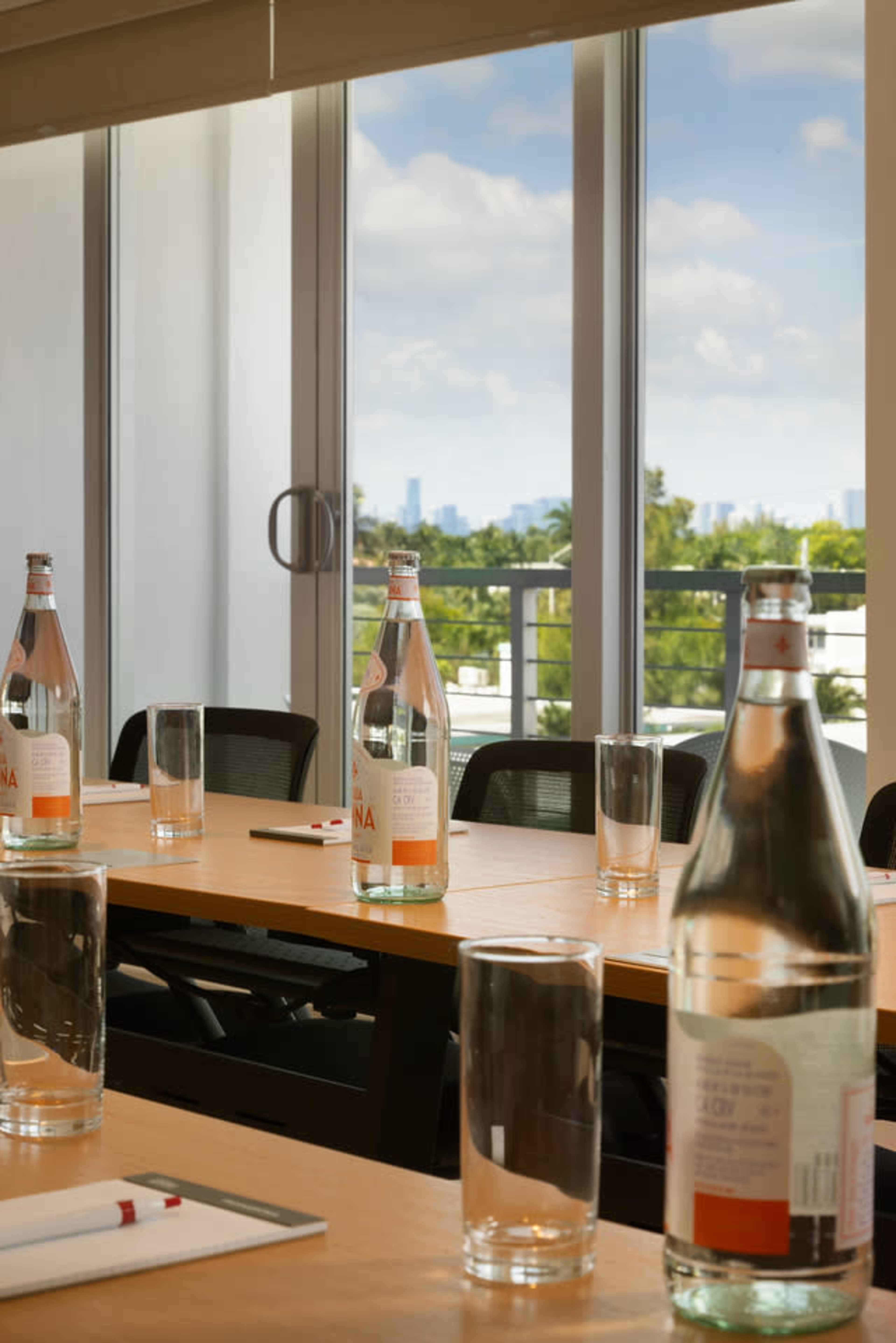 A conference room table is set with several glass bottles of water and empty glasses, with large windows offering a view of a city skyline.